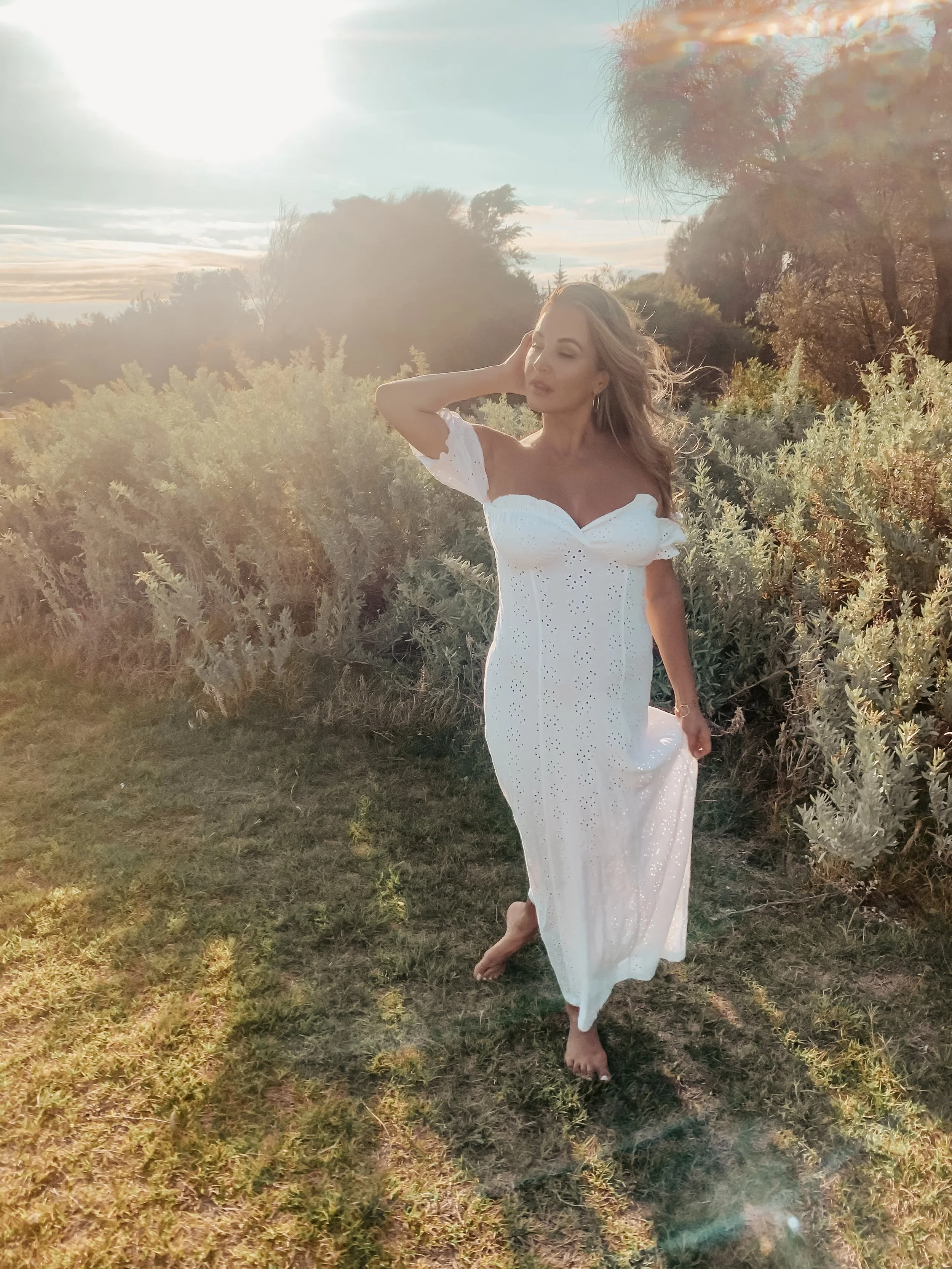 Woman in a white dress walking barefoot outdoors during sunset, with greenery and trees in the background.