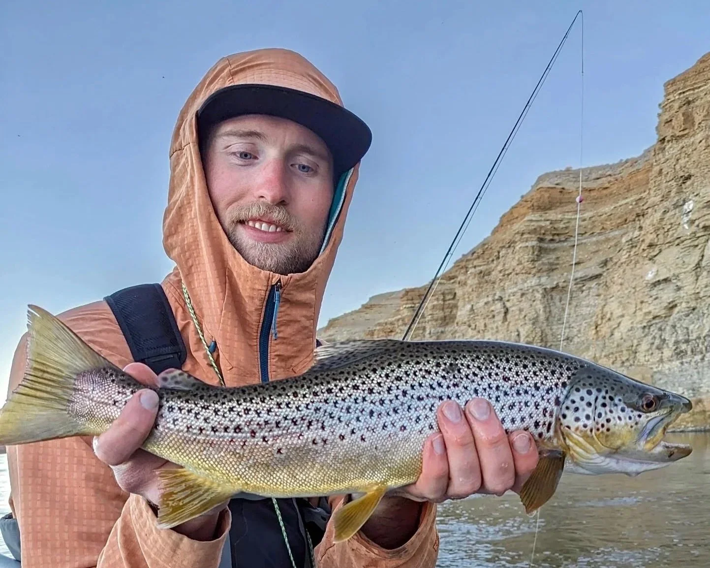 A man wearing a brown hooded jacket and a black cap holding a large brown trout fish with a rocky cliff in the background.