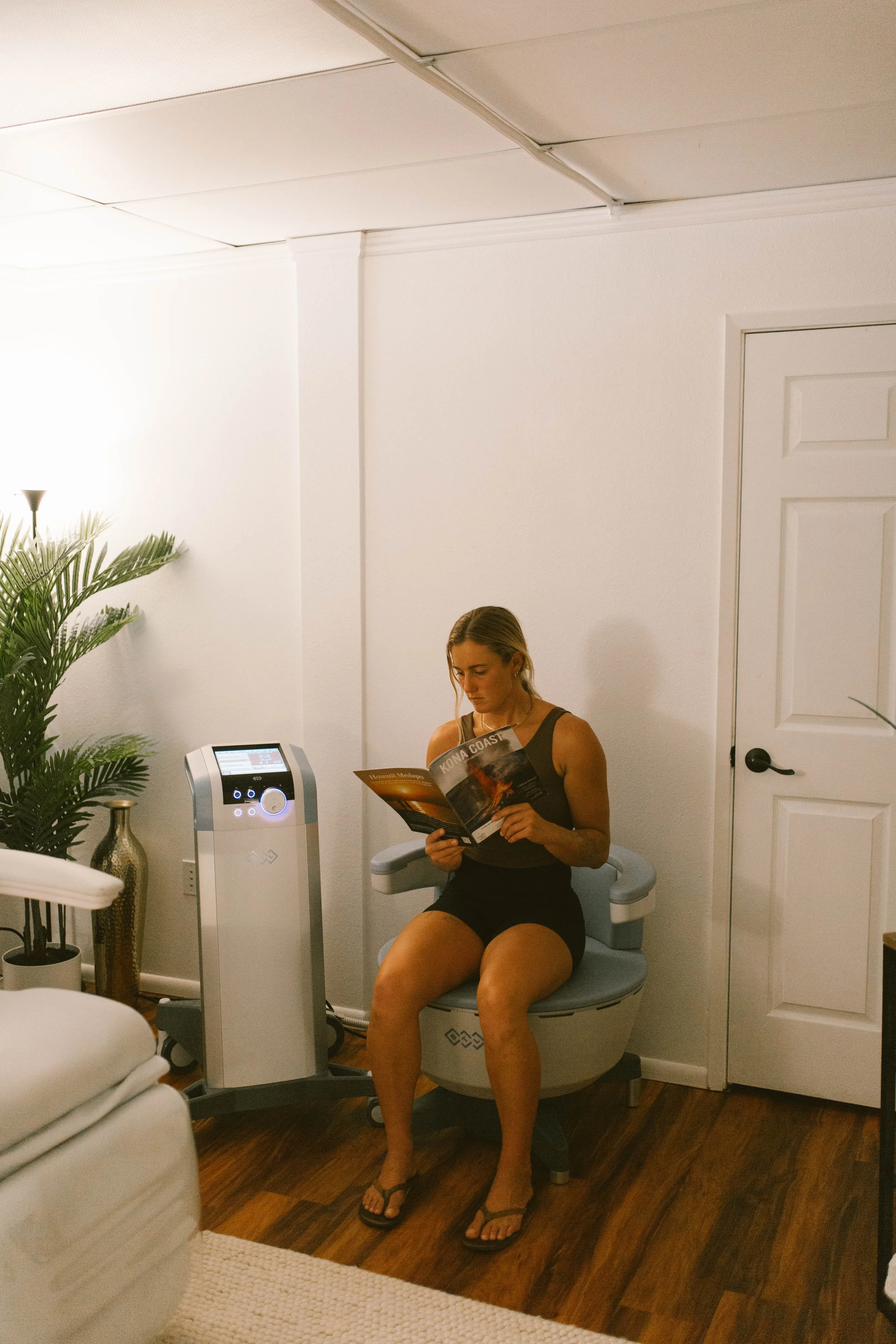 A woman sitting on a toilet reading a magazine in a small, well-lit room.