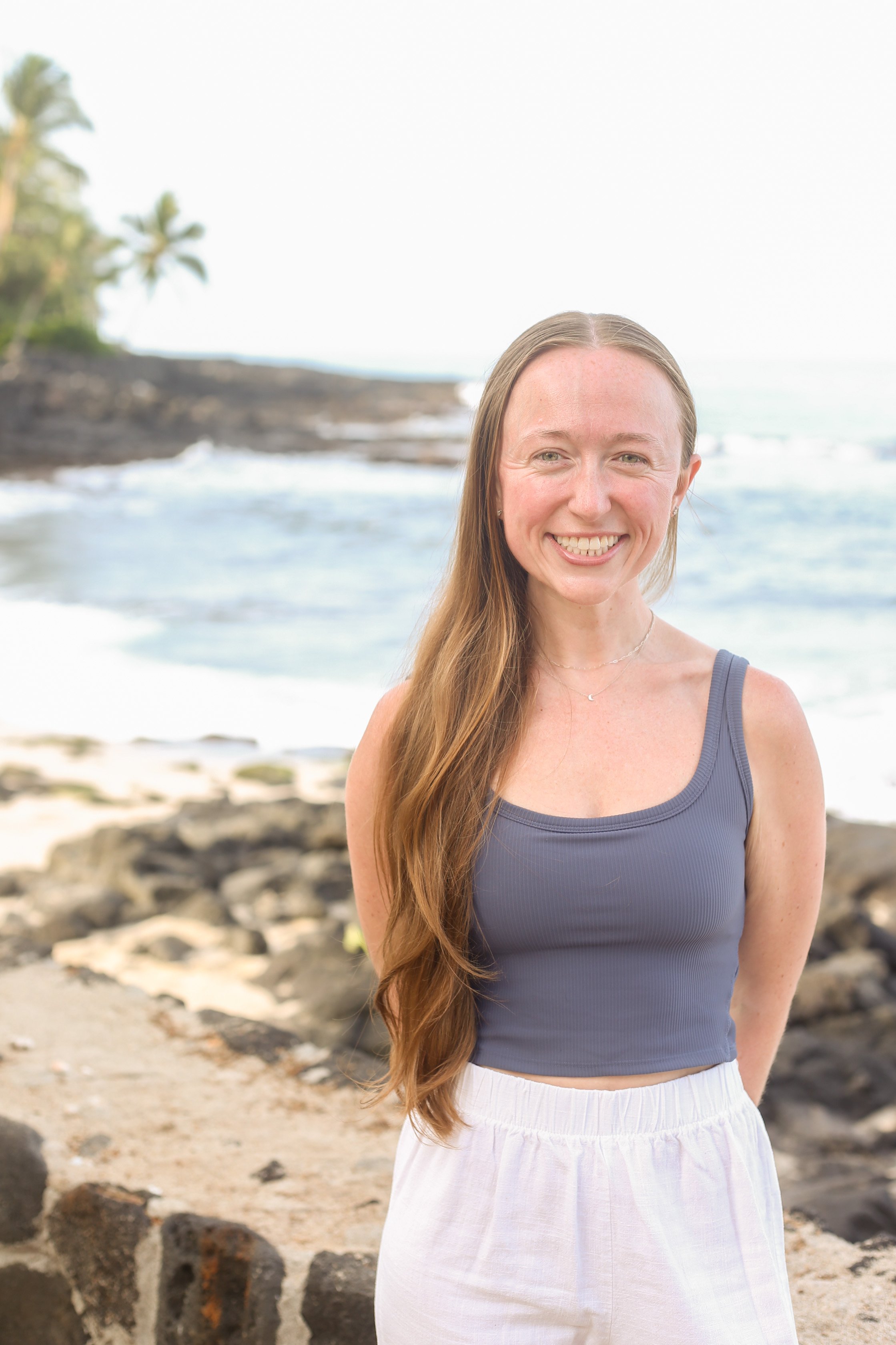 A women with long wavy hair smiling at the camera on a beach with rocks and ocean in the background.