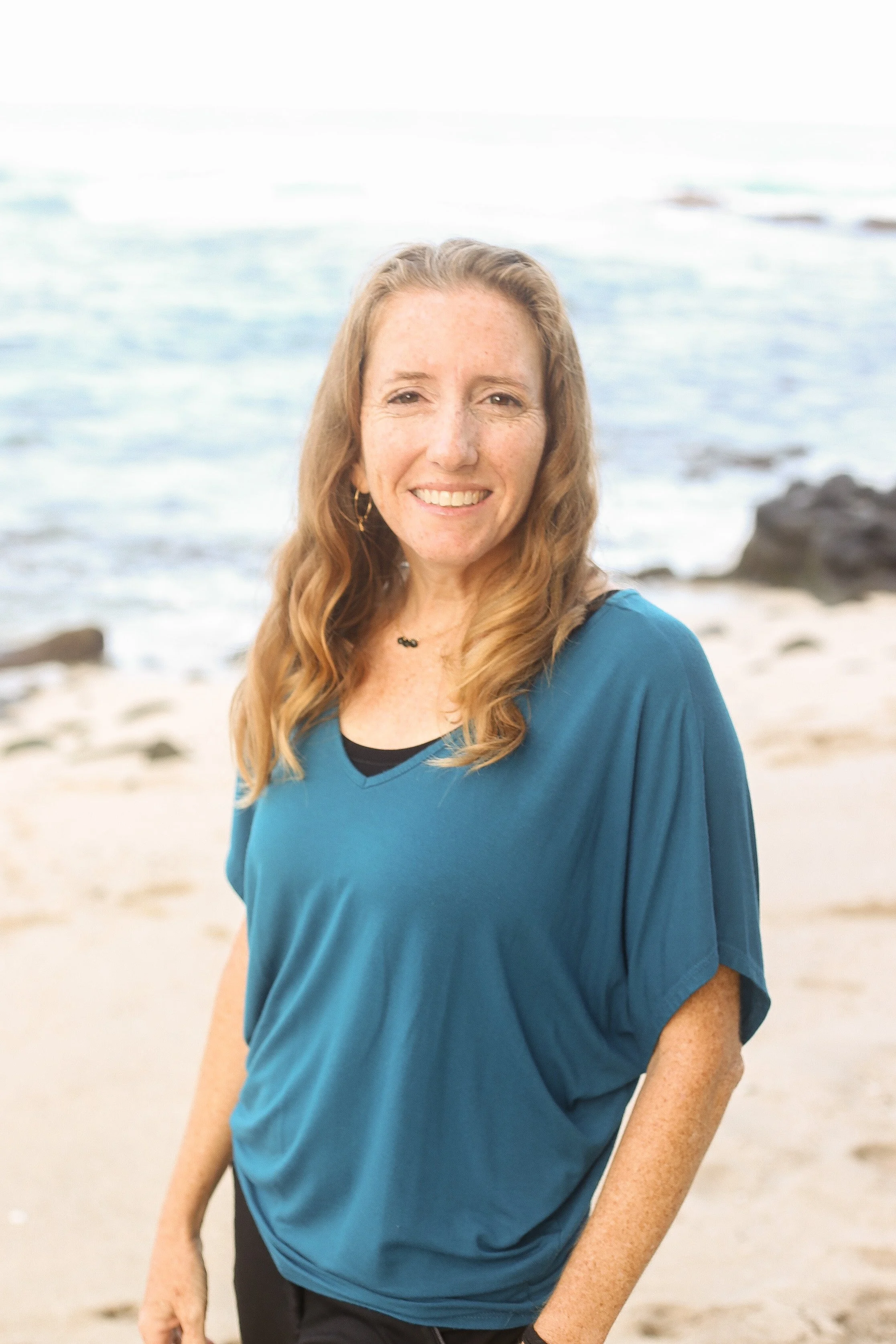 A woman with long wavy red hair, smiling, standing on a sandy beach near the water, wearing a teal shirt.