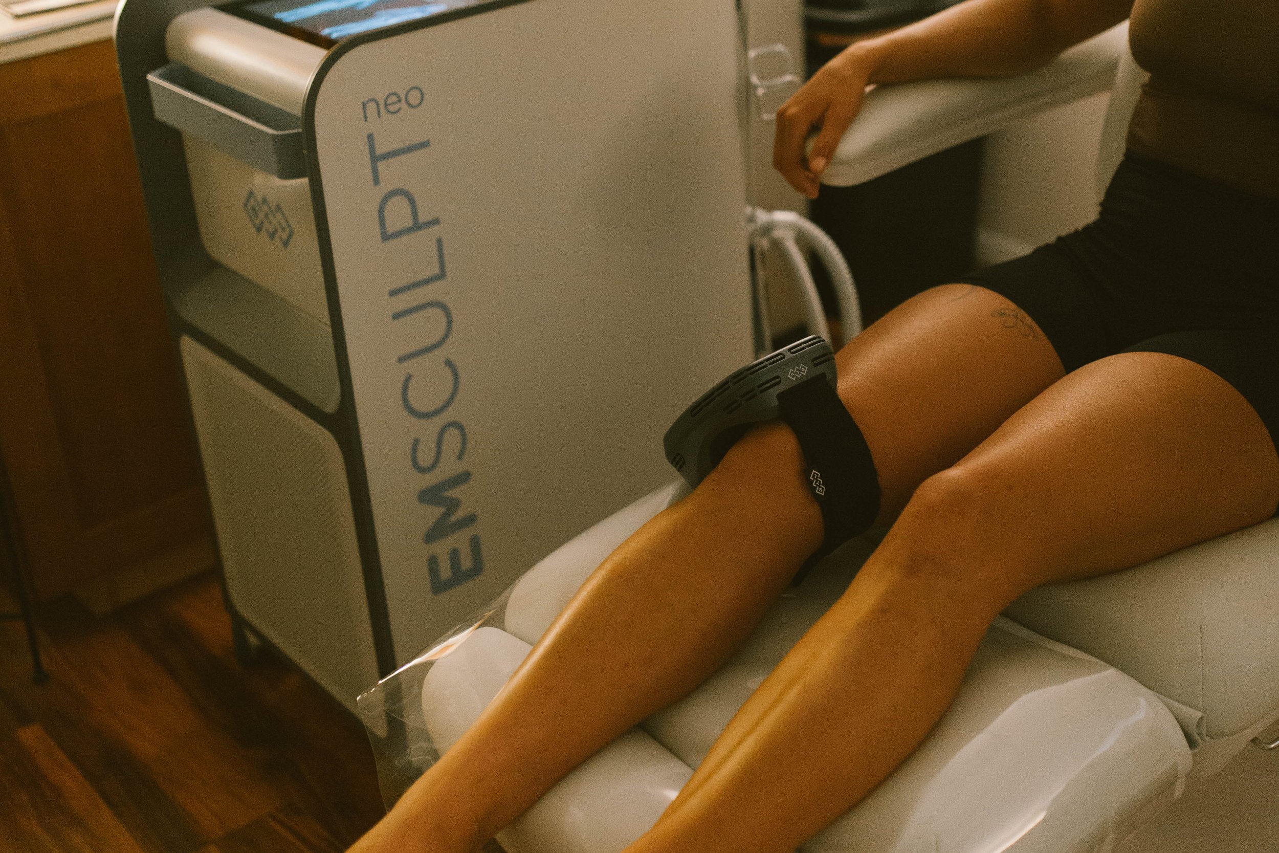 Person lying on a medical examination table with a blood pressure cuff around their arm, with an emsculpt machine nearby.