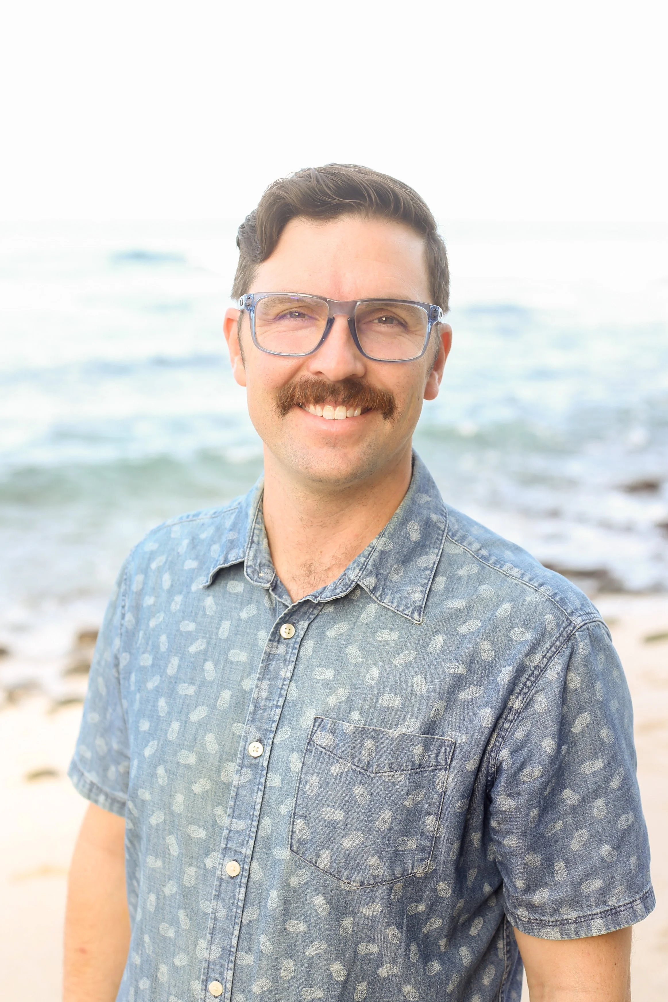 A man with glasses and a mustache smiling at the camera, standing on a beach with the ocean in the background.
