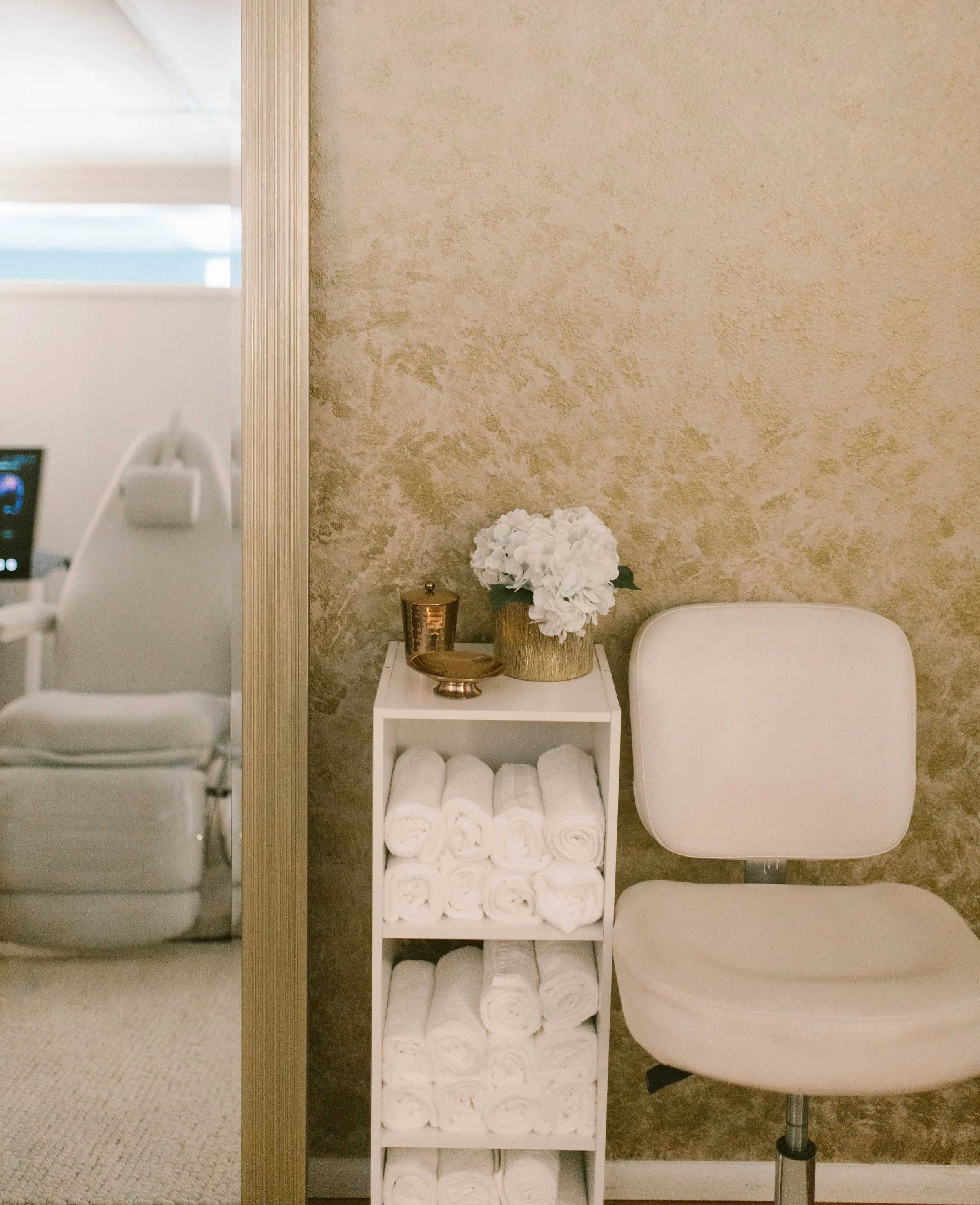 A narrow shelving unit with neatly rolled white towels, a small bronze vase with white flowers, and a bronze cup, placed next to a white chair, against a textured beige wall in a spa or medical office setting.