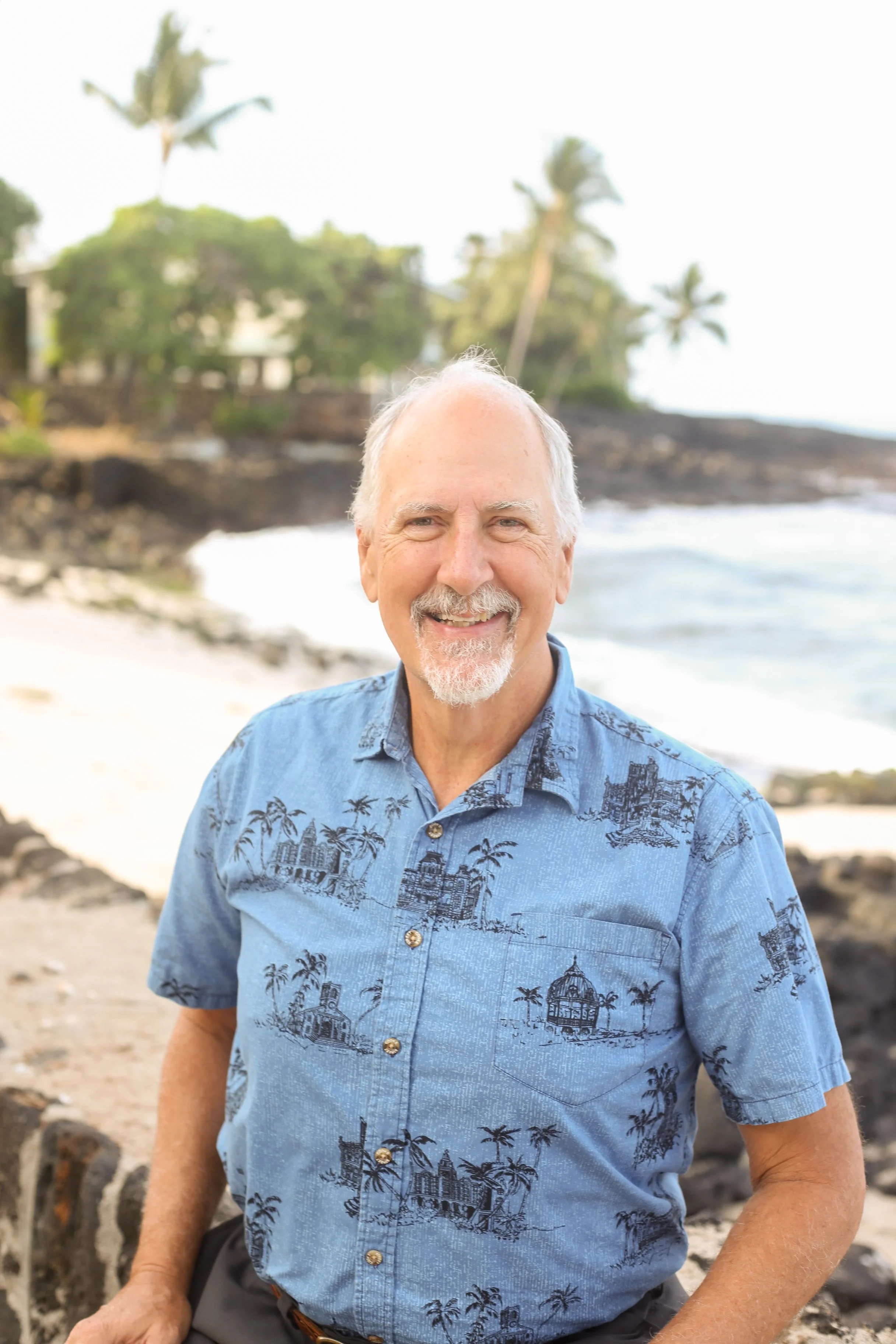 A smiling older man with a white beard and mustache, wearing a blue Hawaiian shirt with palm tree and building patterns, stands on a beach with palm trees and ocean waves in the background.