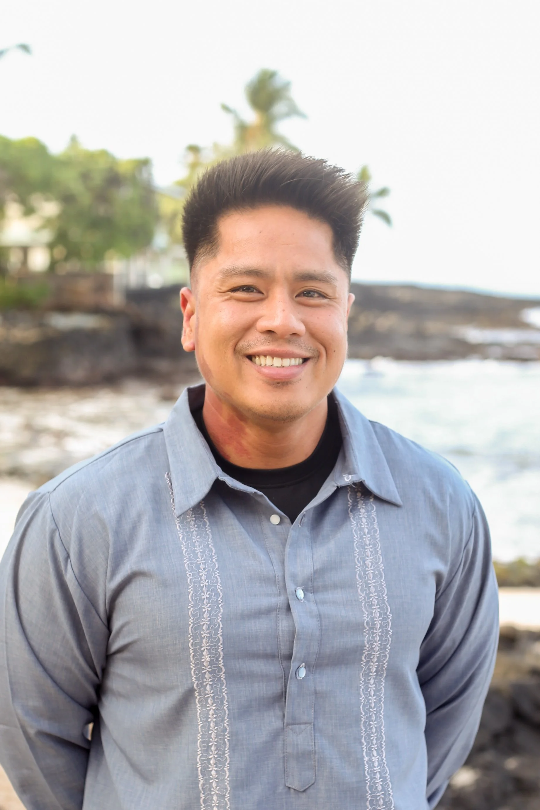 A smiling man standing outdoors with a beach and palm trees in the background.