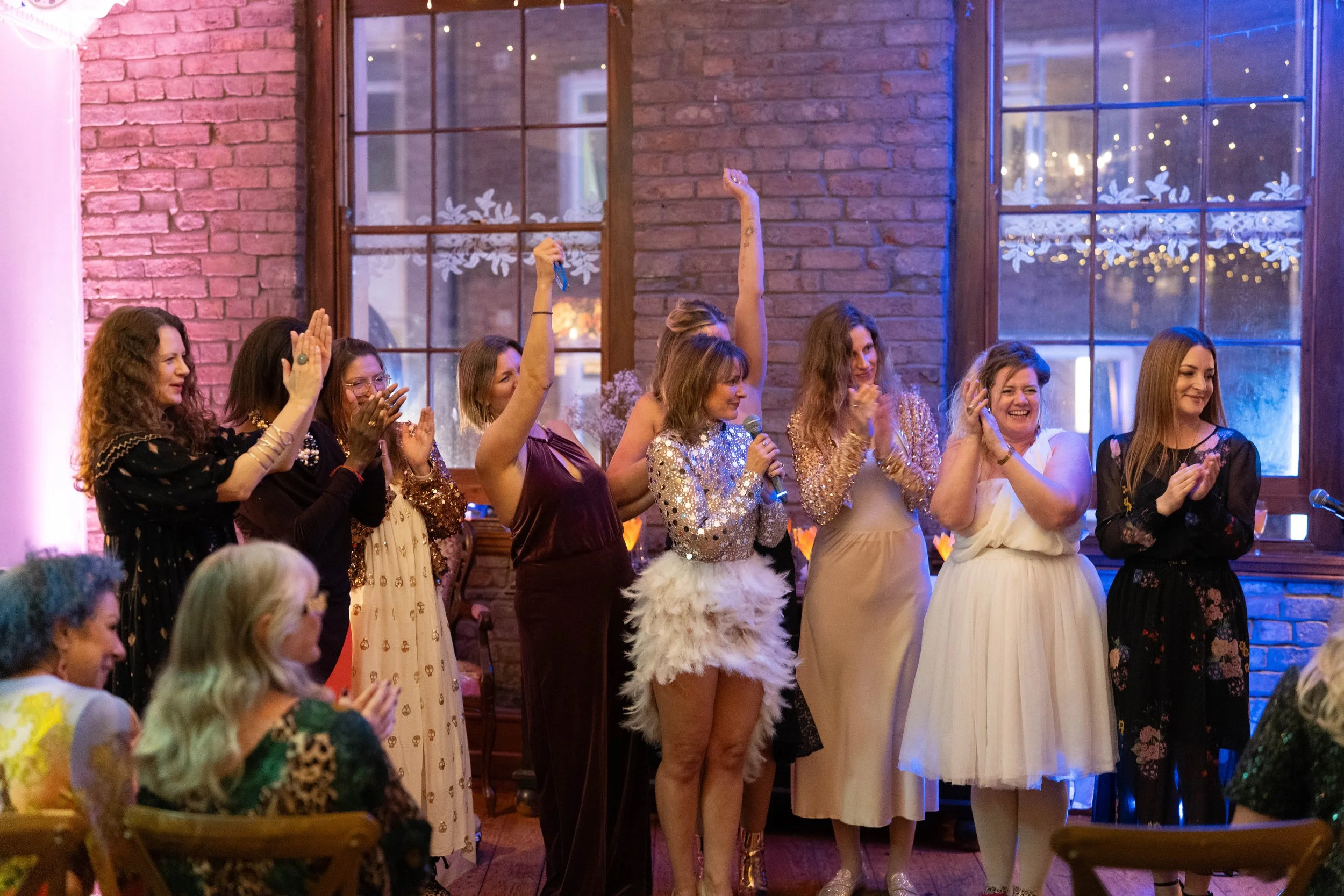 A group of women on stage at a celebration, some clapping, one holding a microphone, one with a raised arm, in a decorated venue with large windows and ambient lighting.