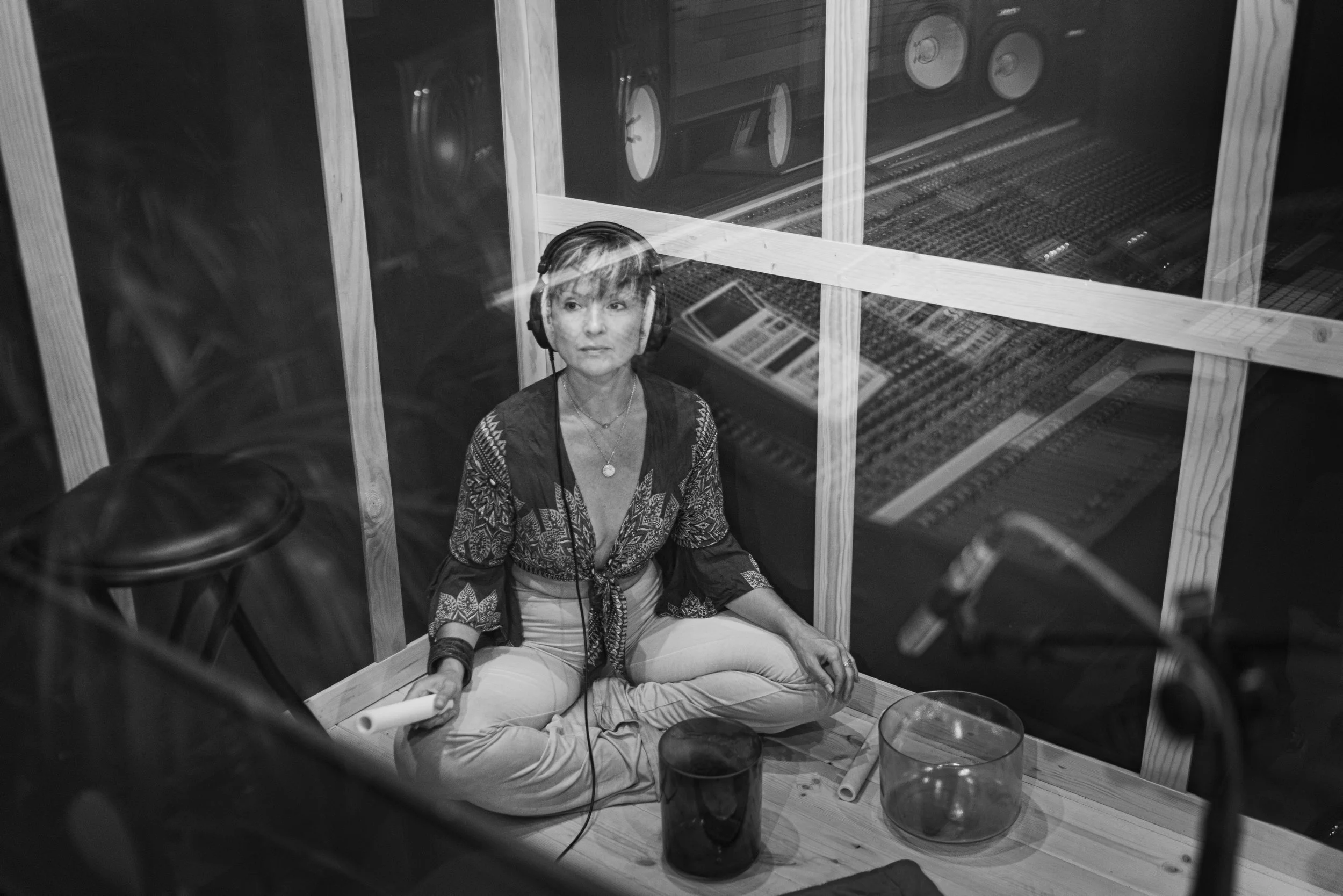 A woman with headphones sitting cross-legged in a recording studio, holding a rolled paper in one hand, with a glass bowl and a container on the floor nearby, seen through a window into the studio.