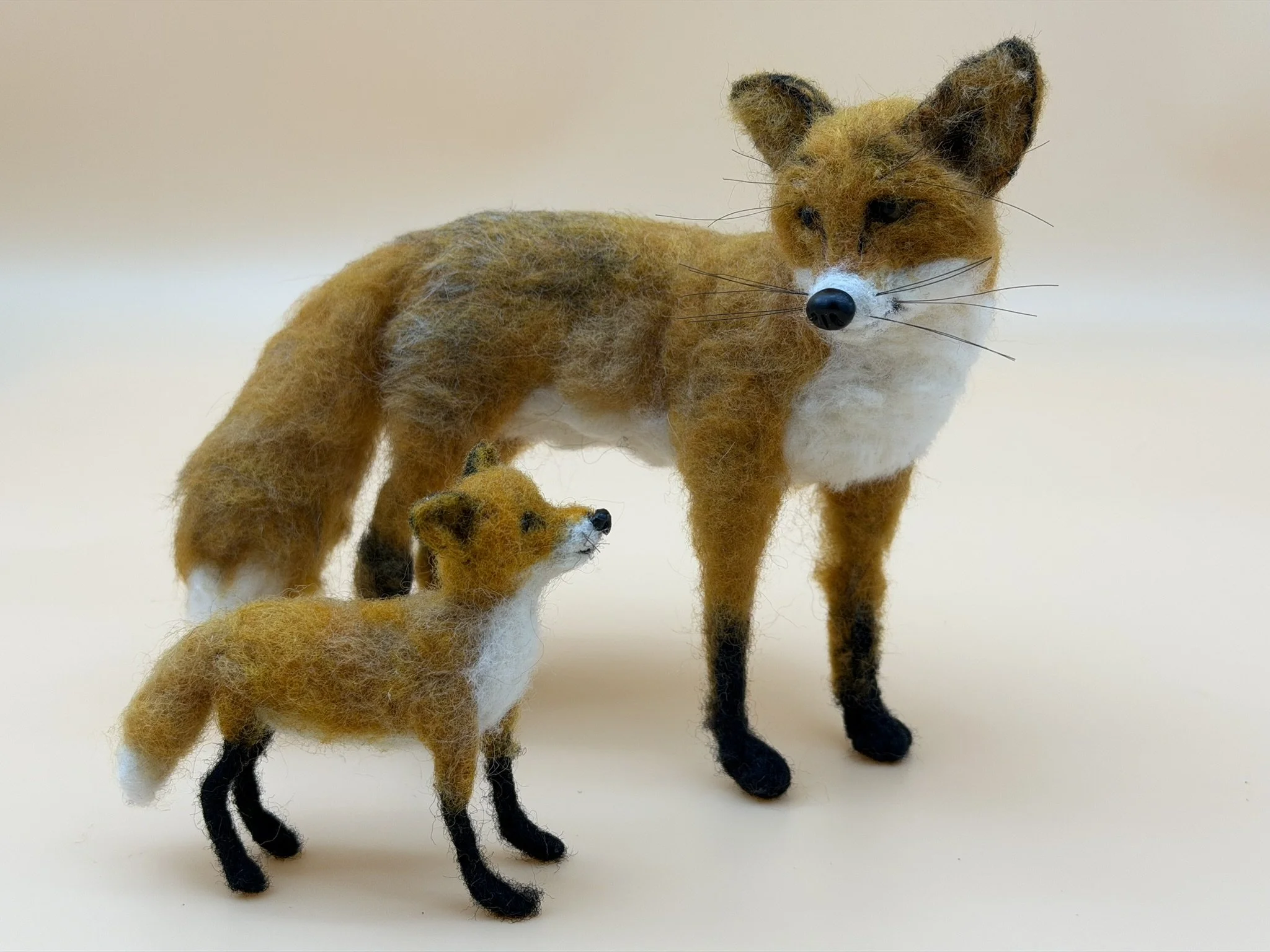 Felted wool sculptures of two foxes, an adult and a cub, standing against a plain light background.