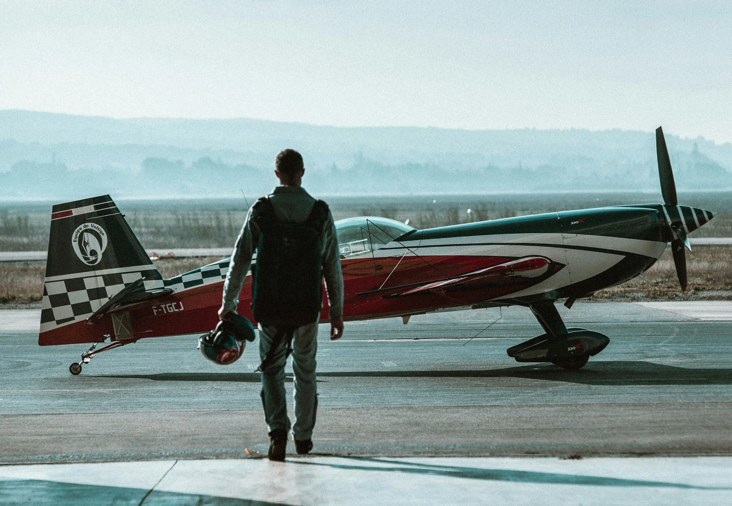 A person walking towards a small modern airplane on the tarmac at an airport, holding a helmet in their right hand, with a vast landscape and distant hills in the background during daytime.