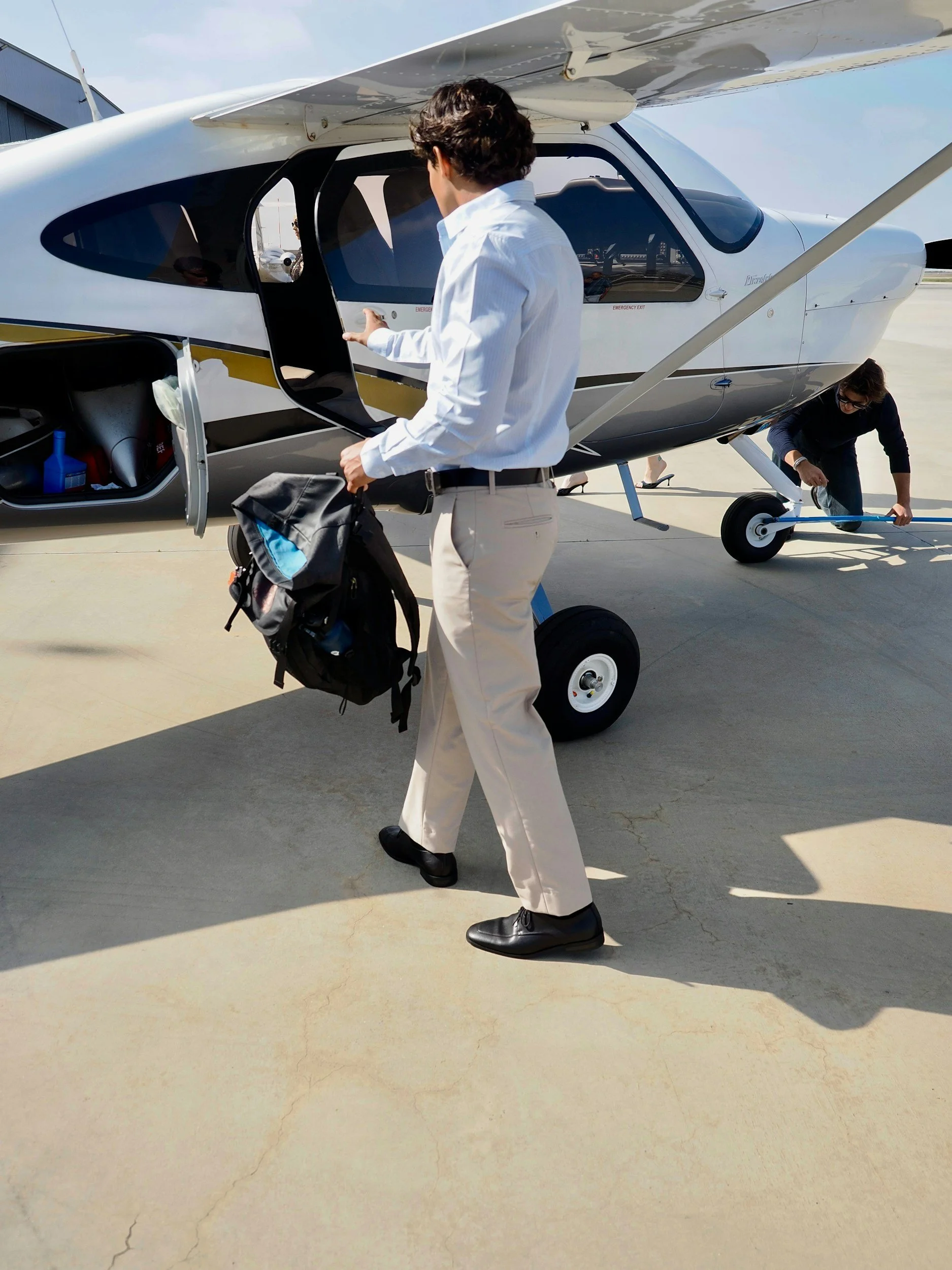 A man in a white shirt and beige pants is walking towards a small aircraft on a tarmac, with a backpack in his hand. Another person is kneeling near the aircraft's wheel, working on it. The aircraft is white with black and gold accents.