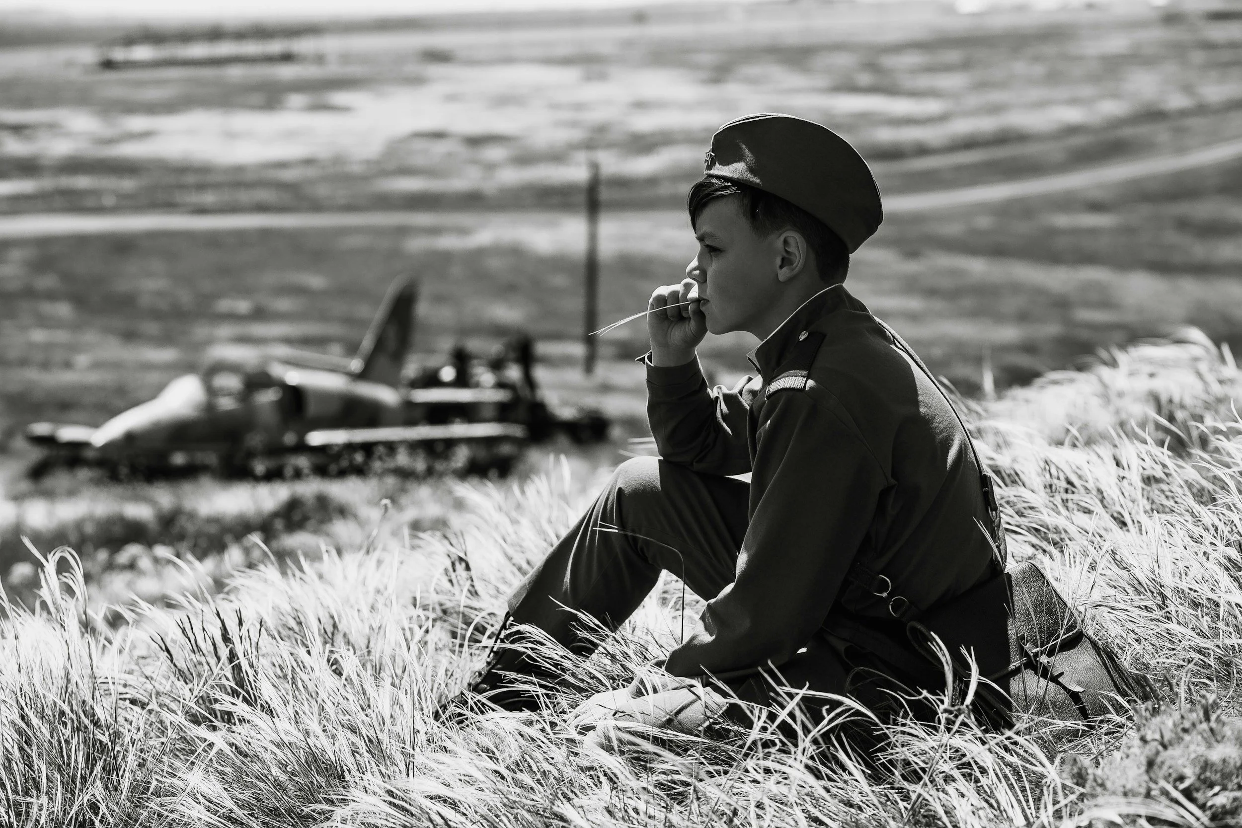 A young soldier in military uniform sitting in a grassy field, smoking a cigarette, with a tank in the background.