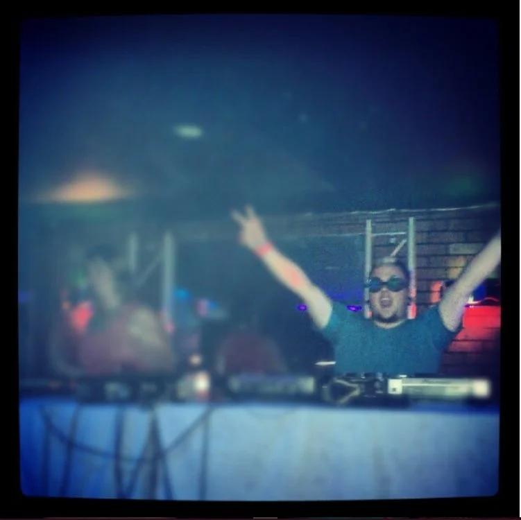 DJ Molex wearing sunglasses and a t-shirt with arms raised behind a DJ booth in a club with colorful lights.