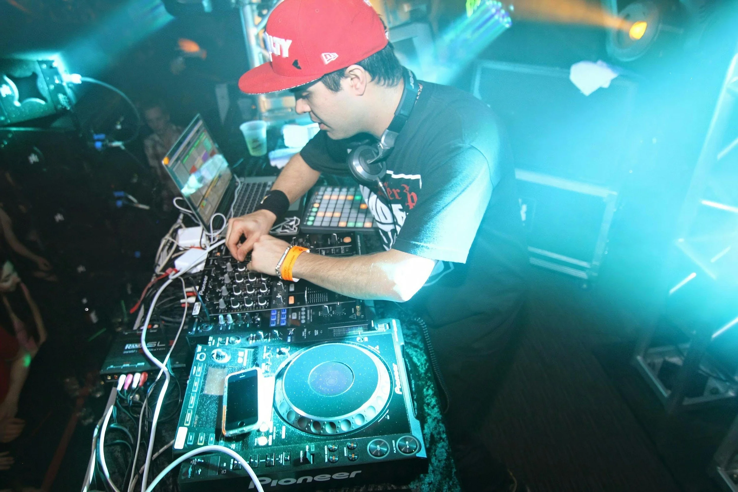 A DJ in a black T-shirt and red cap performing at a nightclub with bright stage lights, surrounded by DJ equipment including a Pioneer mixer and a laptop.