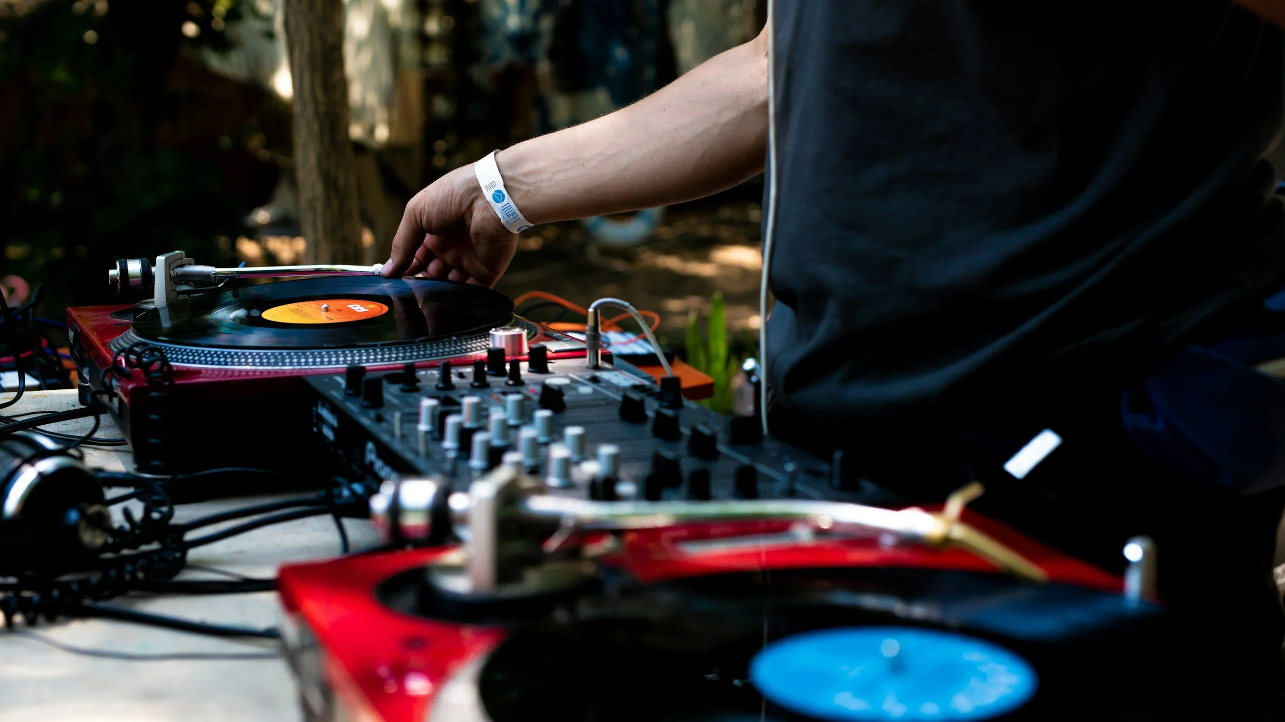 A DJ playing music outdoors with turntables and mixer on a table, hand adjusting a record on the turntable, blurred trees in the background.
