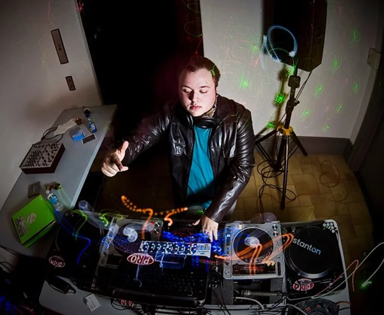 DJ Molex wearing a leather jacket and headphones around his neck is performing at a party, using mixing equipment under colorful lights.