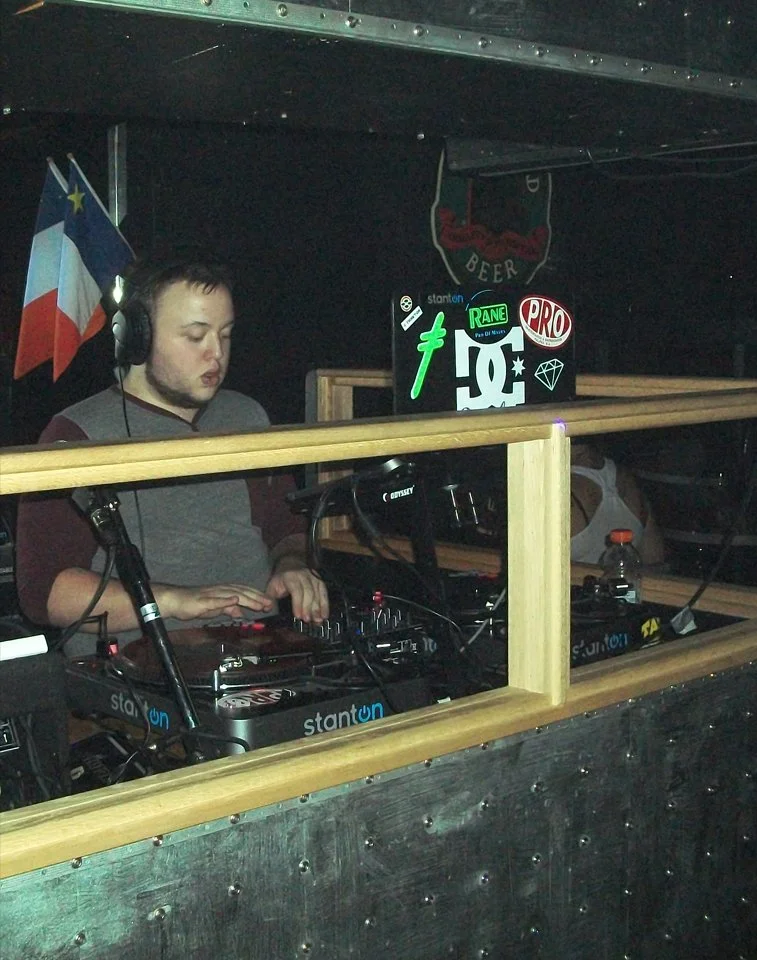 DJ Molex wearing headphones, mixing music on DJ equipment behind a wooden barrier at a club or event, with flags and stickers on the wall in the background.