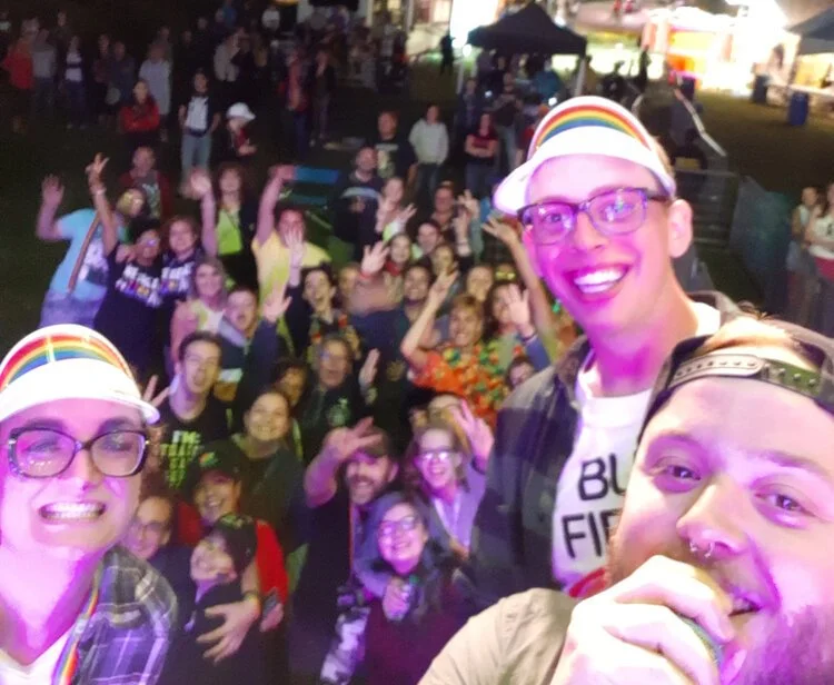 A group of diverse people at an outdoor event at night, smiling and waving at the camera, with some wearing rainbow hats and glasses, suggesting a celebration or festival.
