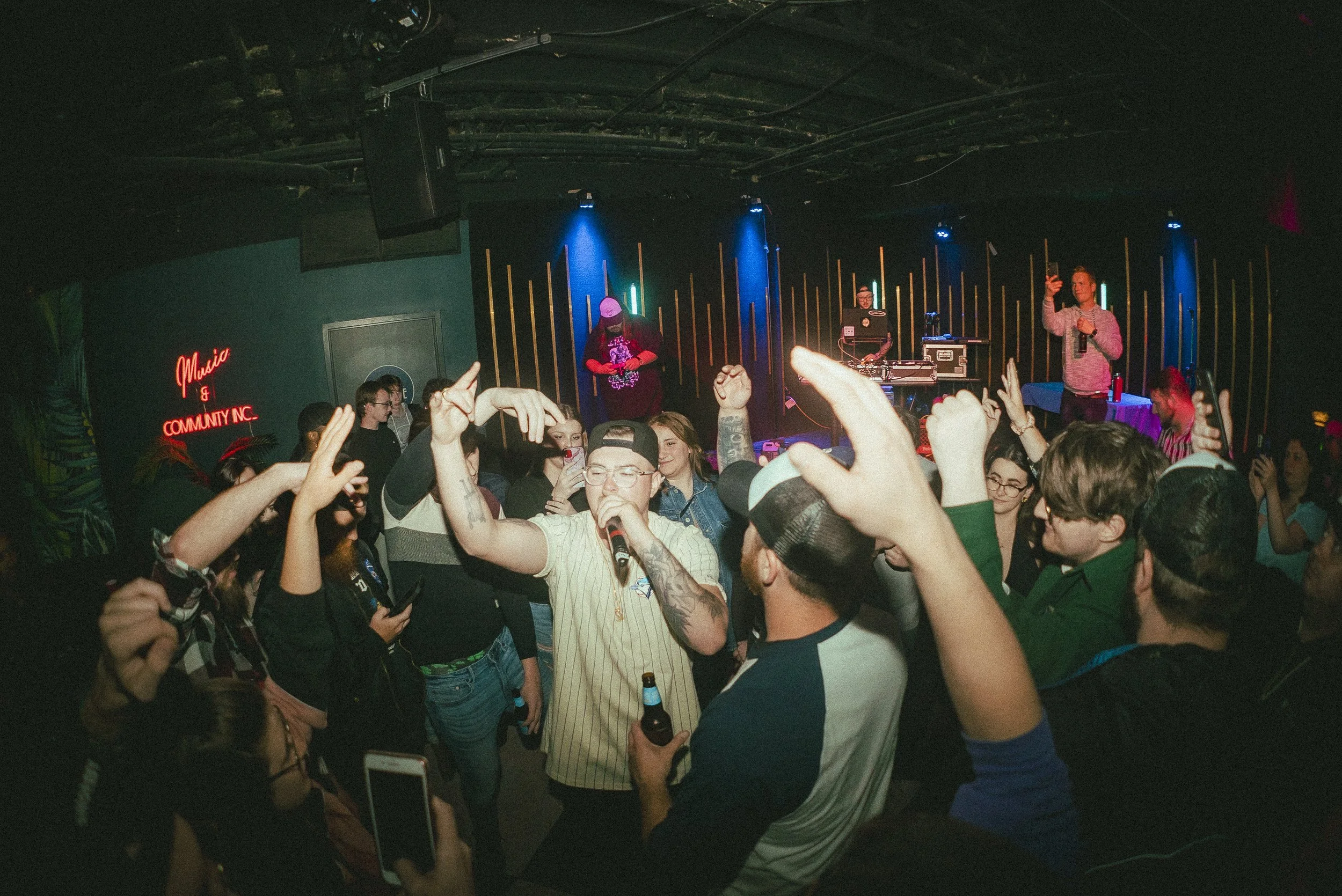 Crowd of people dancing and singing at a concert or club with a DJ on stage and live music performance, neon signs, and colorful lights.