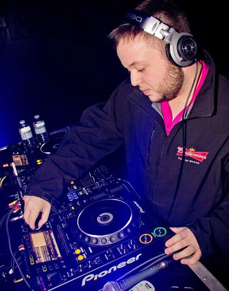 DJ Molex wearing headphones, operating a Pioneer DJ controller in a dark setting with bottles and equipment in background.