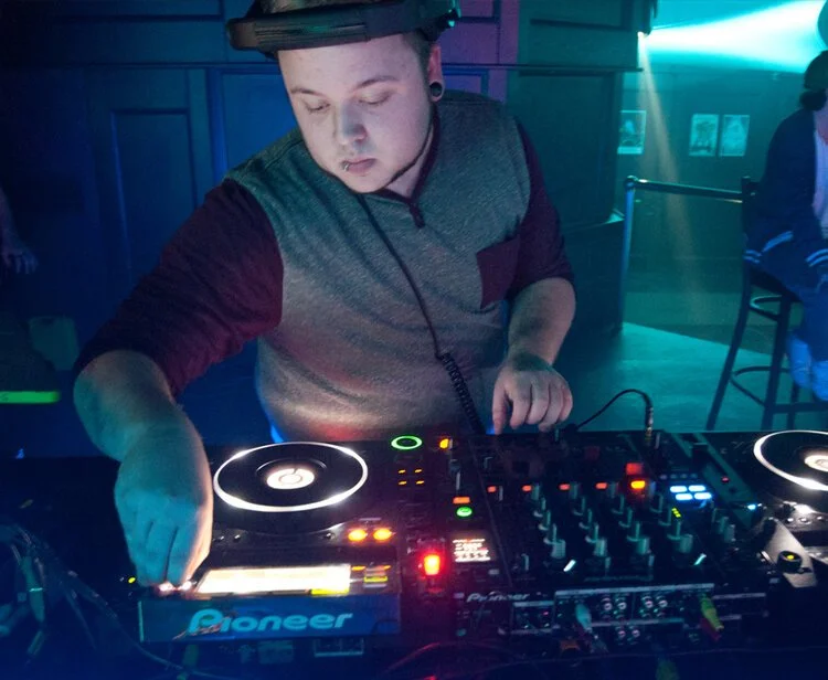  DJ Molex wearing a gray and maroon long sleeve shirt mixing music at a club, with colorful lighting and a Pioneer DJ controller.