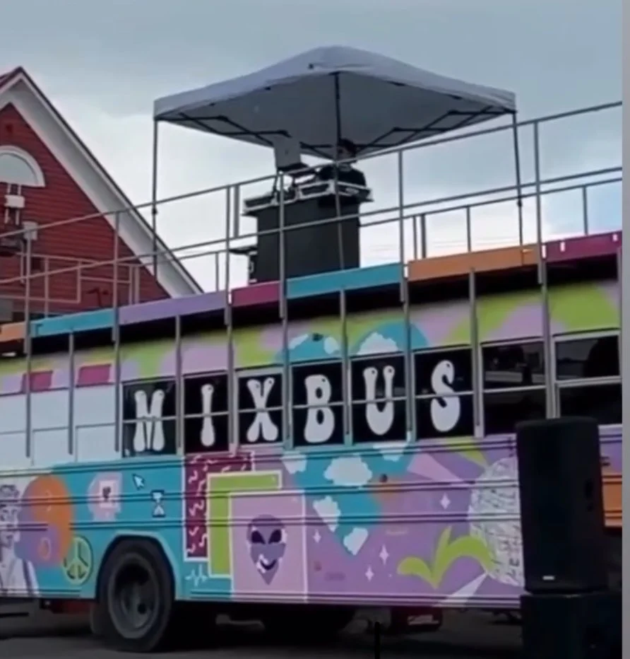 DJ Molex performing on top of a colorful bus with decorations and the word 'MIXBUS' in the windows, on a city street with a building and overcast sky in the background.