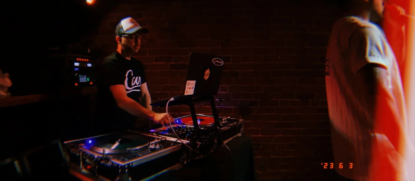 DJ Molex playing music at a set of turntables and a mixer, with a brick wall background, in a dimly lit venue.