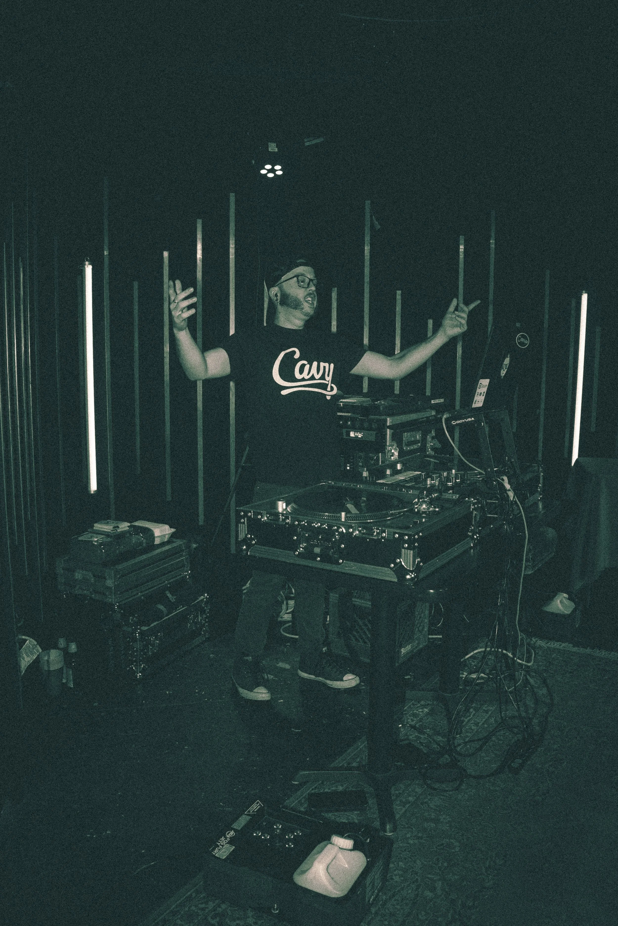 DJ Molex wearing a black t-shirt with the word 'Cavy' in white is performing at a dark venue with black walls and vertical light strips, using DJ equipment on a table.