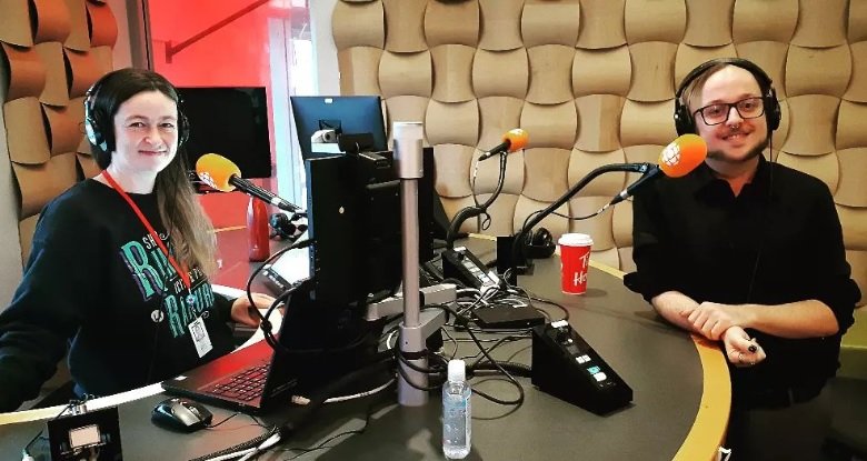 Two people in a radio studio wearing headphones, sitting at a table with microphones, computers, and audio equipment.