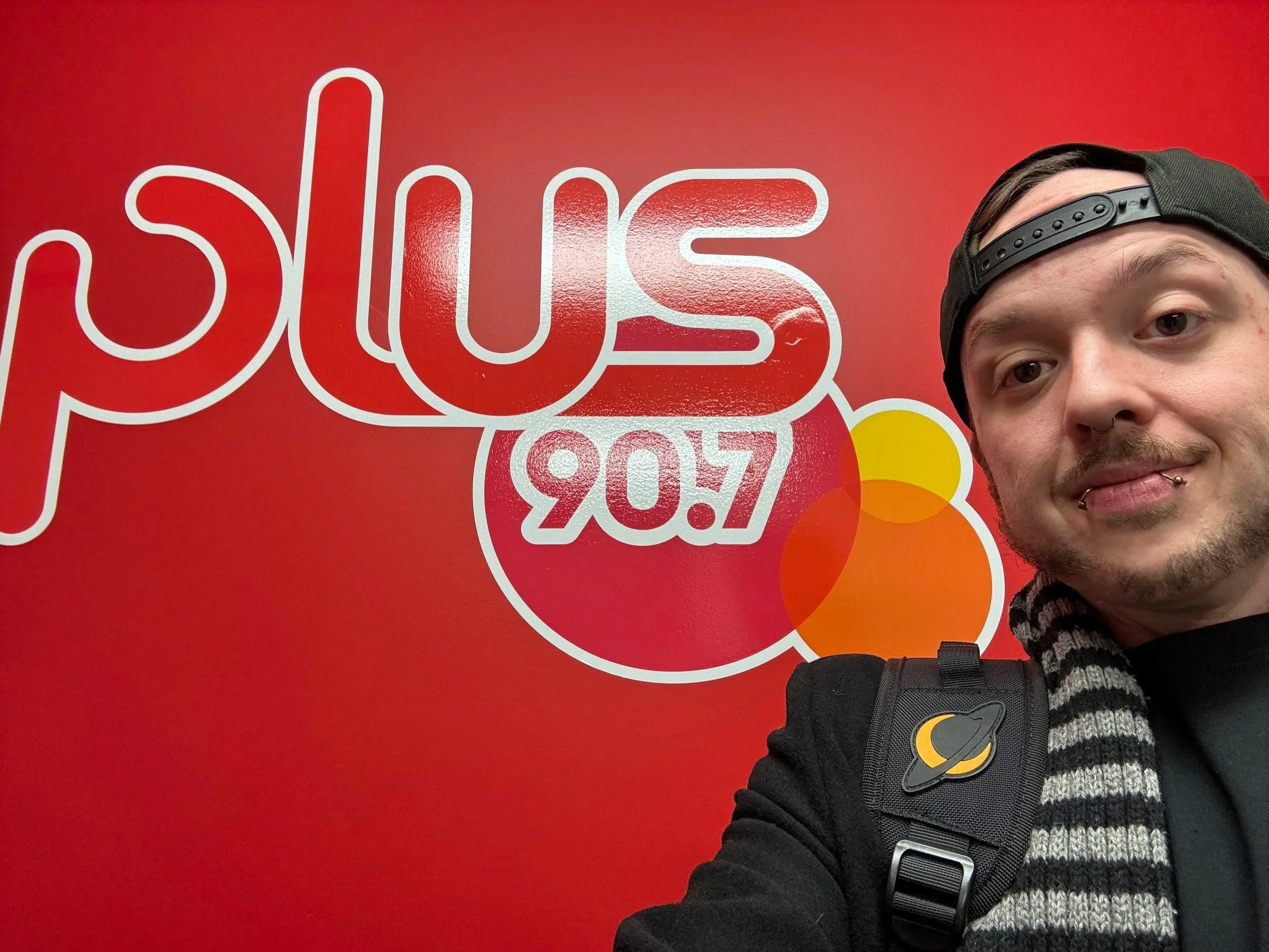 Close-up of DJ Molex, taking a selfie in front of a bright red wall with the Plus 90.7FM logo.