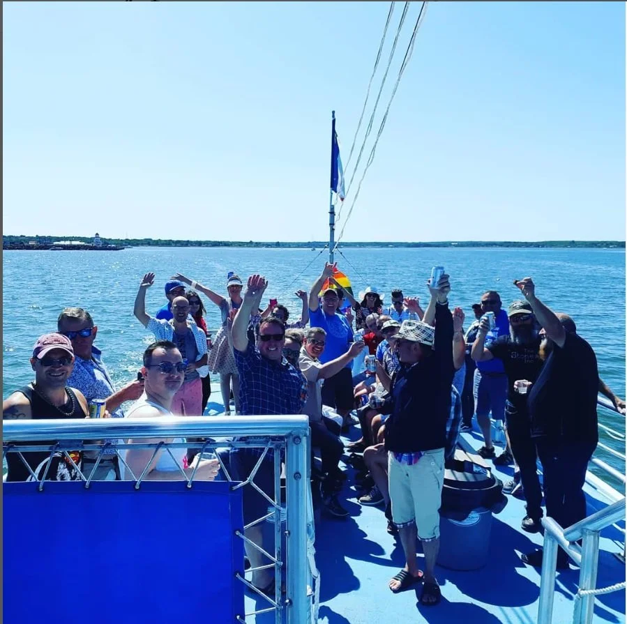 Group of people on a boat enjoying sunny weather, some waving and relaxed, with the water and distant shoreline in the background.