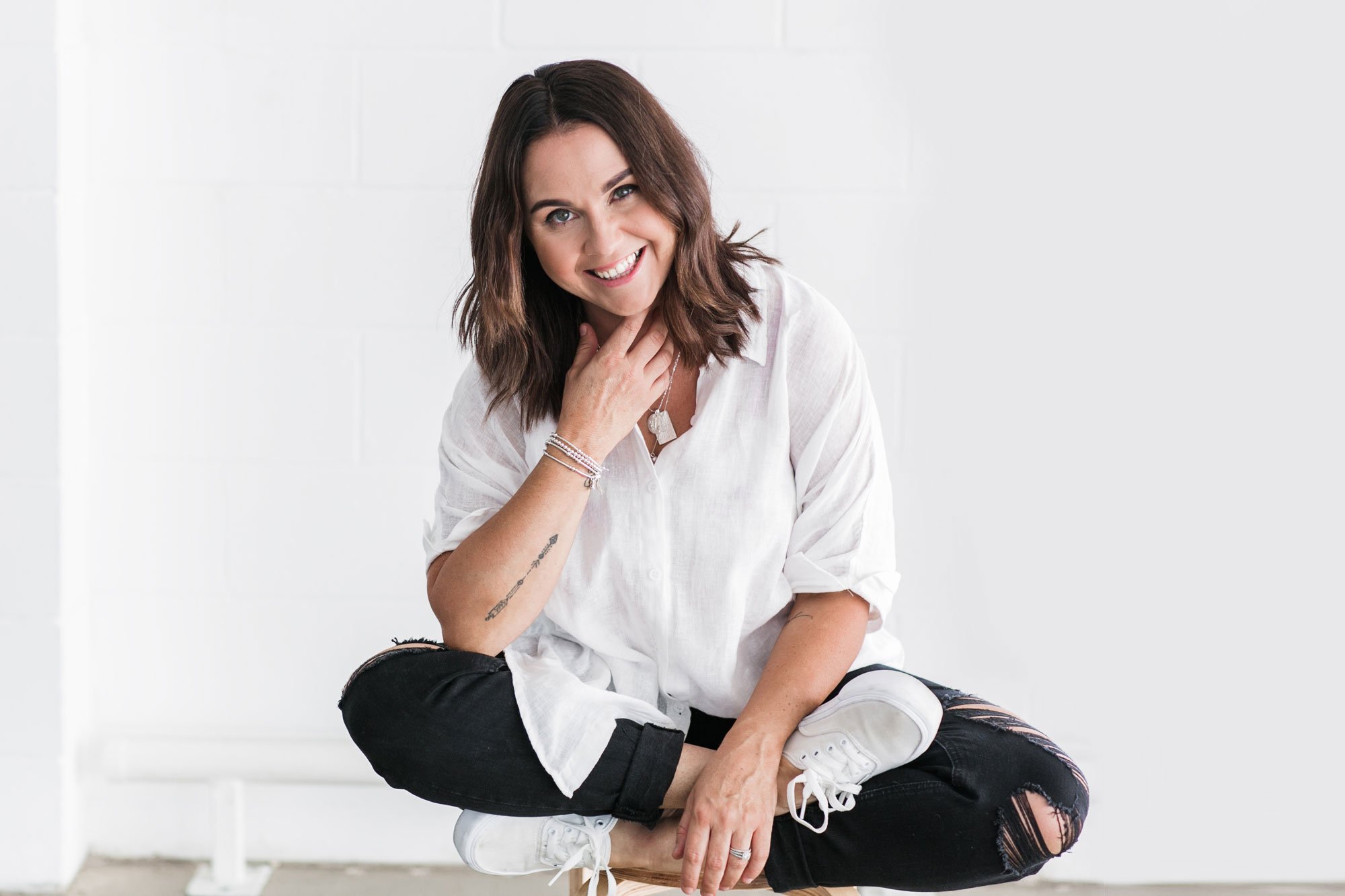 Shanny sitting on a wooden stool in a warehouse, wearing a black and white outfit and smiling at the camera