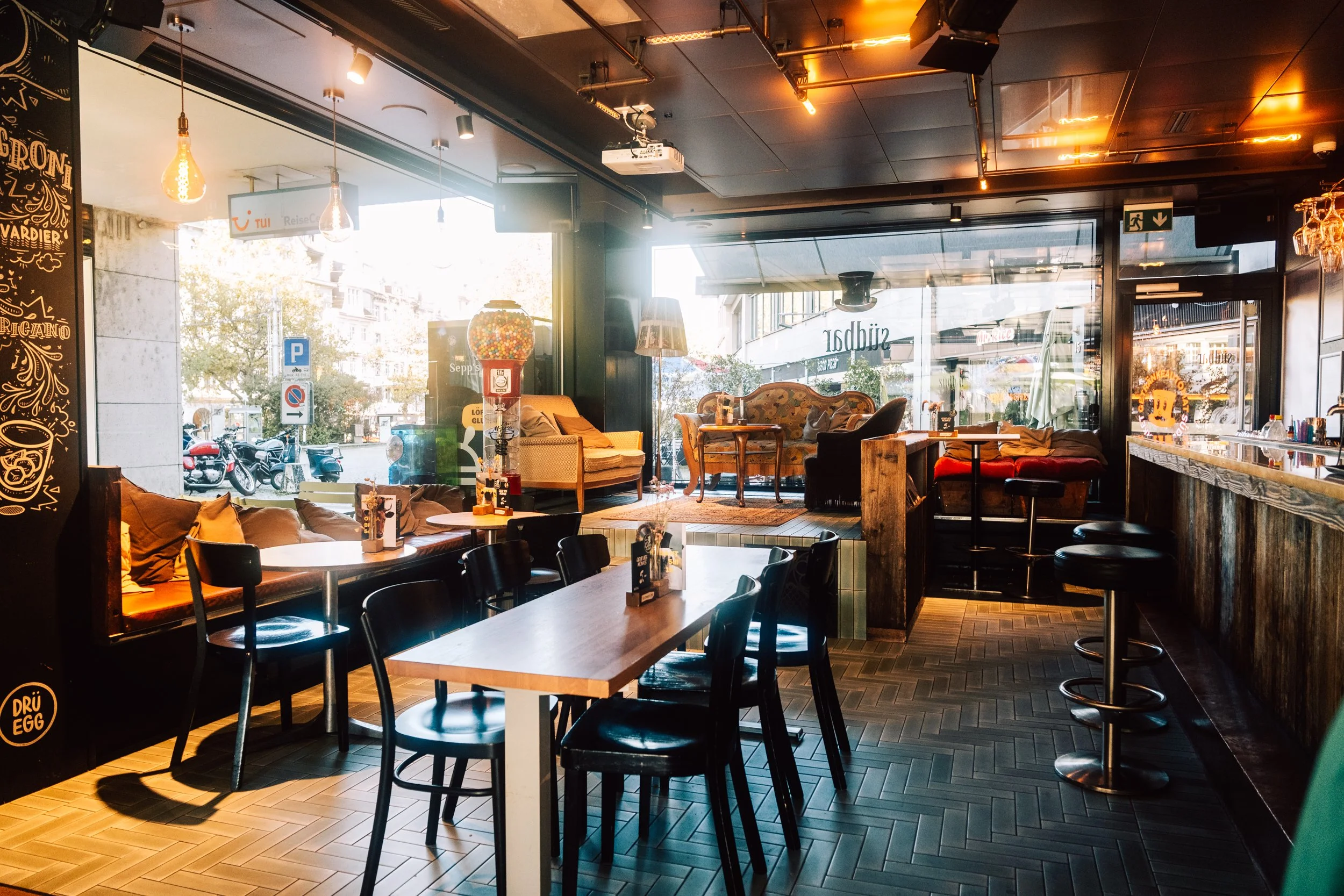 Innenraum eines Cafés mit Tischen, Stühlen, Lounge-Sitzen und großen Fenstern, durch die Straßenverkehr sichtbar ist, bei Tageslicht.