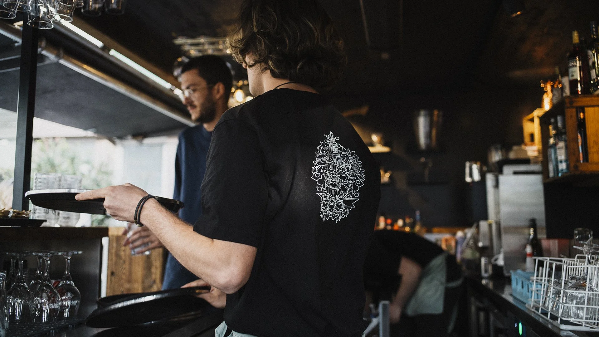 Menschen arbeiten in einer Bar oder Küche, einer trägt ein schwarzes T-Shirt mit einem weißen grafischen Design auf dem Rücken, der andere ist im Hintergrund.