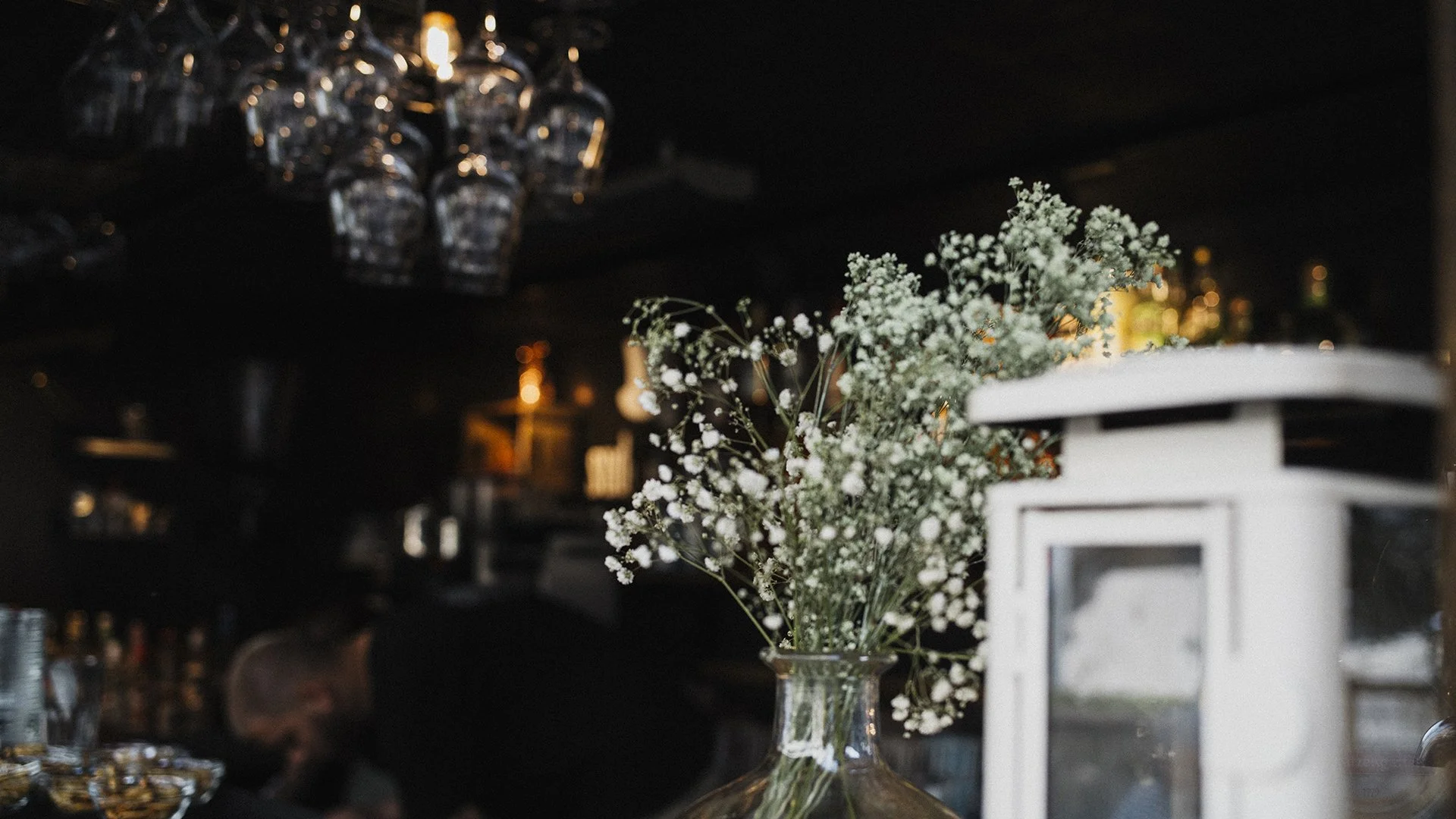 Blumen in einer Vase auf einem Bar-Tresen, im Hintergrund eine dunkle Bar mit Gläsern und Flaschen, und eine Person, die an der Bar arbeitet.