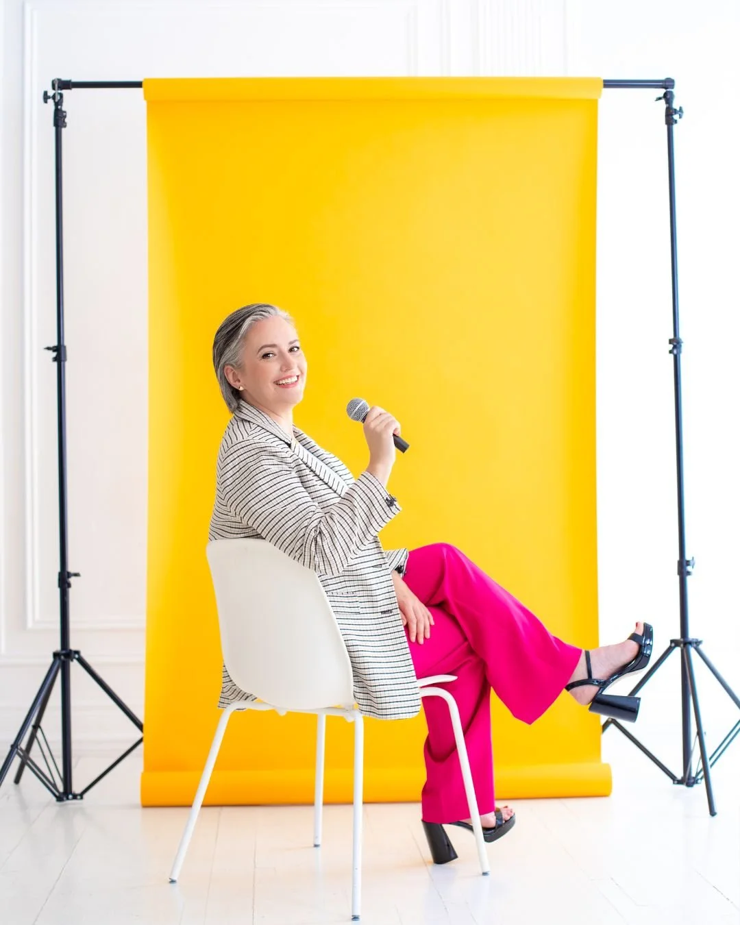Female light skinned keynote speaker with salt and pepper hair, seated on a white chair holding a microphone, smiling confidently in front of a bright yellow backdrop during a professional speaking photoshoot in Canada.