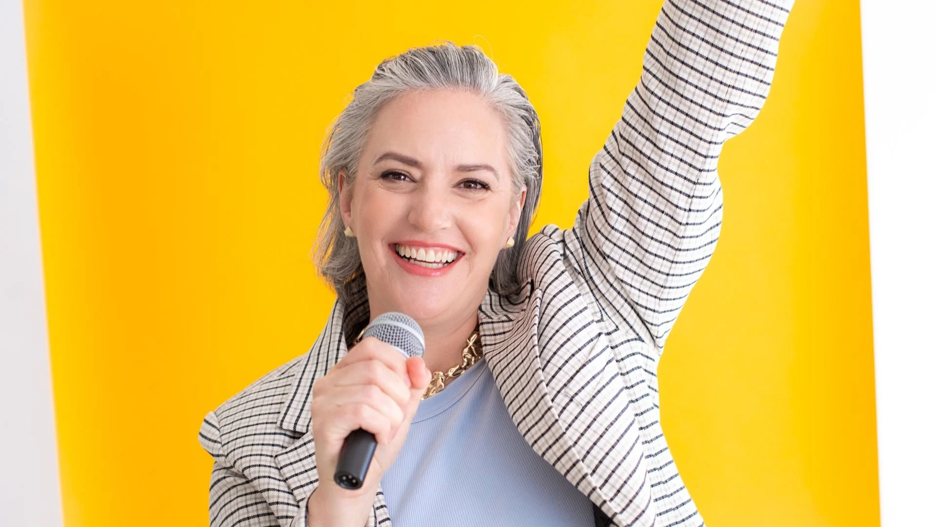 Female light skinned keynote speaker with salt and pepper hair, holding a microphone, smiling confidently in front of a bright yellow backdrop during a professional speaking photoshoot in Canada.