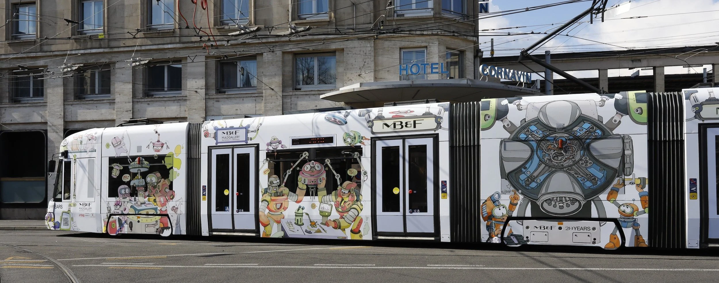 Un tramway recouvert de dessins colorés de robots et de personnages mécaniques, circulant en ville avec un bâtiment d'hôtel en arrière-plan.