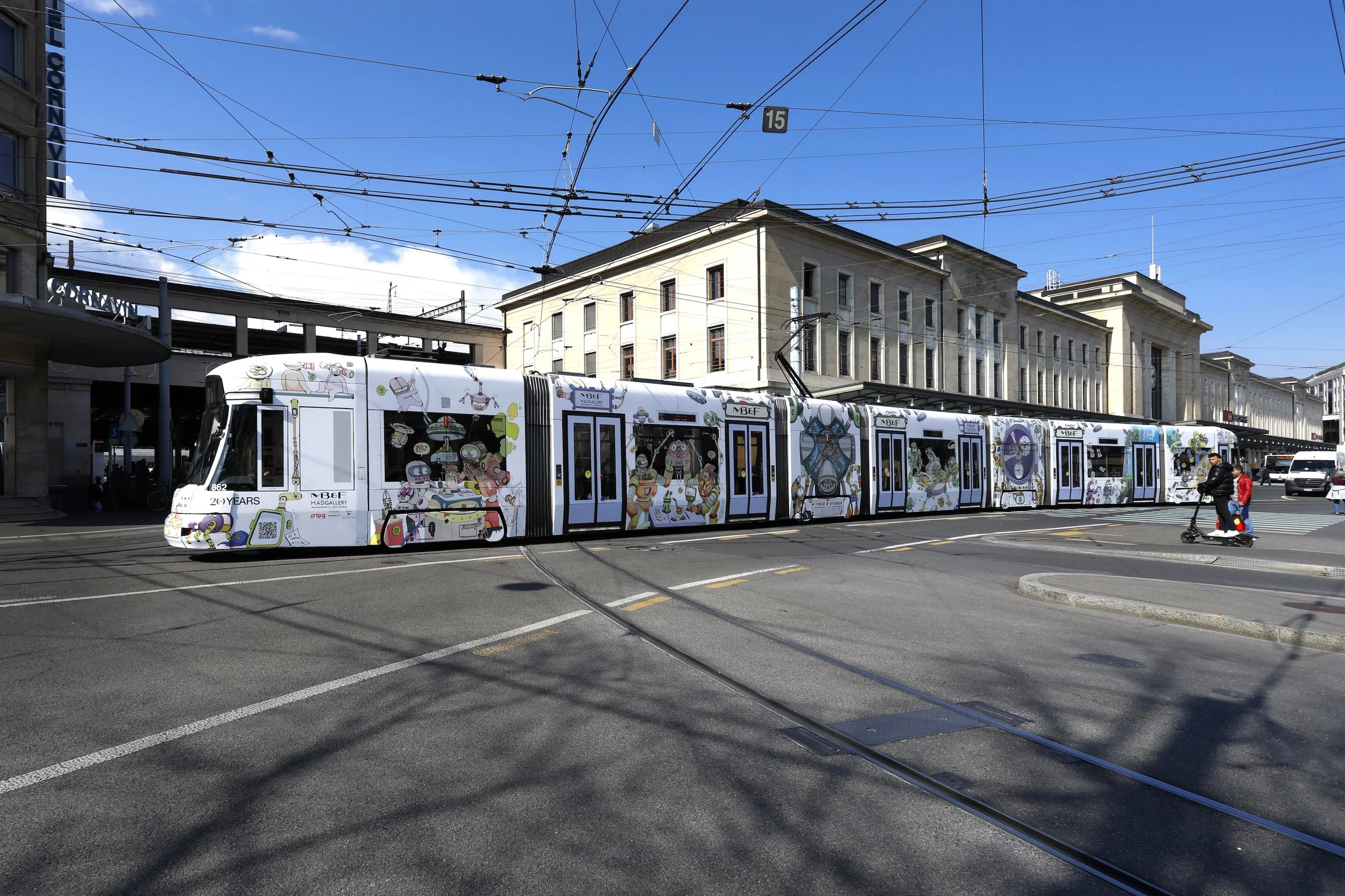 Un tramway décoré avec des illustrations colorées de personnages et d'objets, stationné sur la voie urbaine devant des bâtiments historiques sous un ciel bleu.