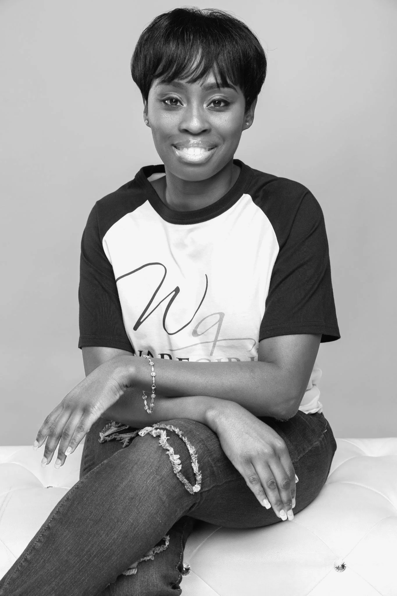 Black and white portrait of a woman with short hair, wearing a casual t-shirt and jeans, sitting on a cushioned surface.