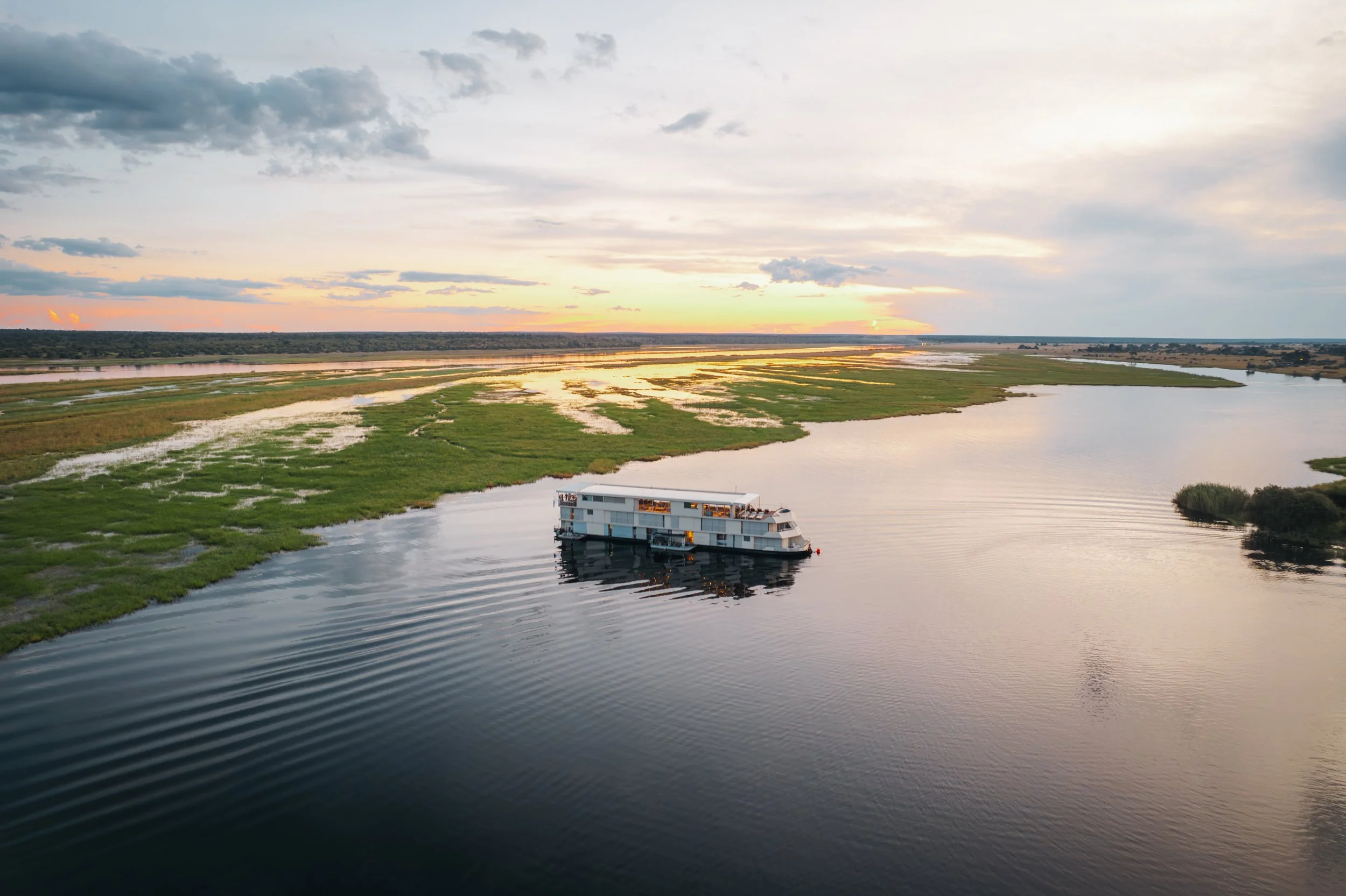 A houseboat floating on a calm river during sunset with green wetlands and cloudy sky in the background.