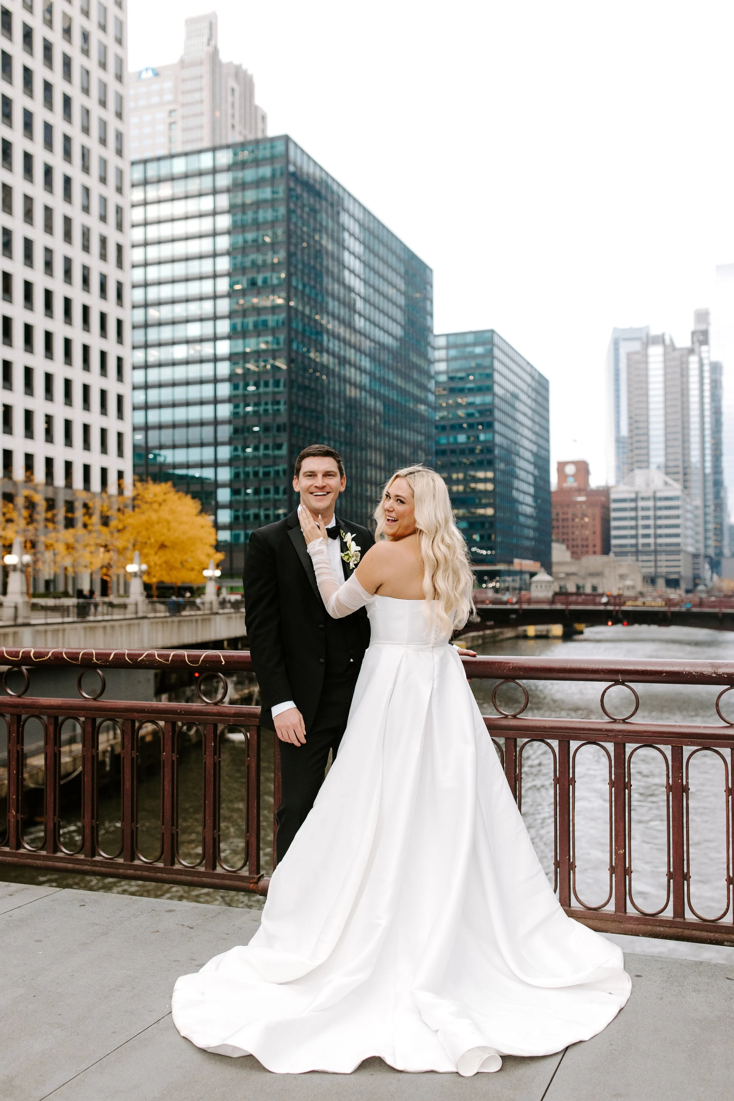 A bride and groom stand together on a bridge in an urban area with tall buildings in the background. The bride wears a strapless white wedding gown with long sheer sleeves, and the groom wears a black tuxedo with a white shirt and tie. They smile happily, sharing a joyful moment.