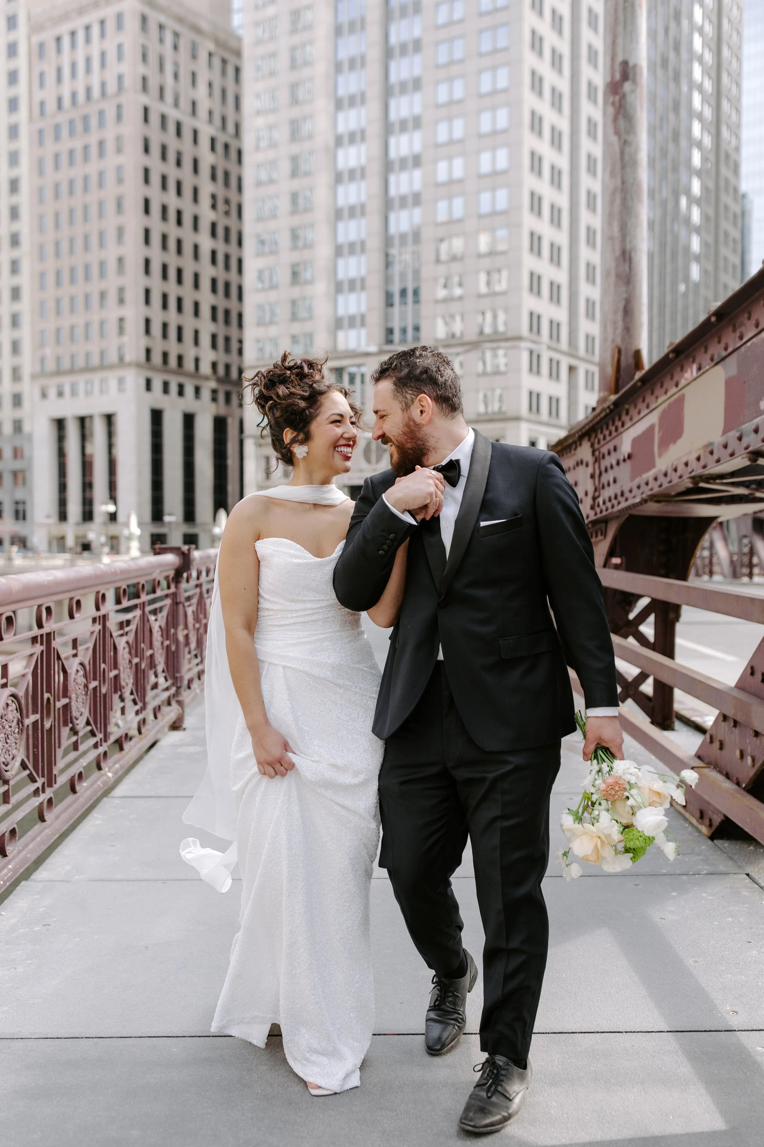 A newlywed couple walking on a city bridge, smiling and gazing at each other, with tall buildings in the background. The bride is wearing a white wedding dress, and the groom is in a black tuxedo holding a bouquet of flowers.