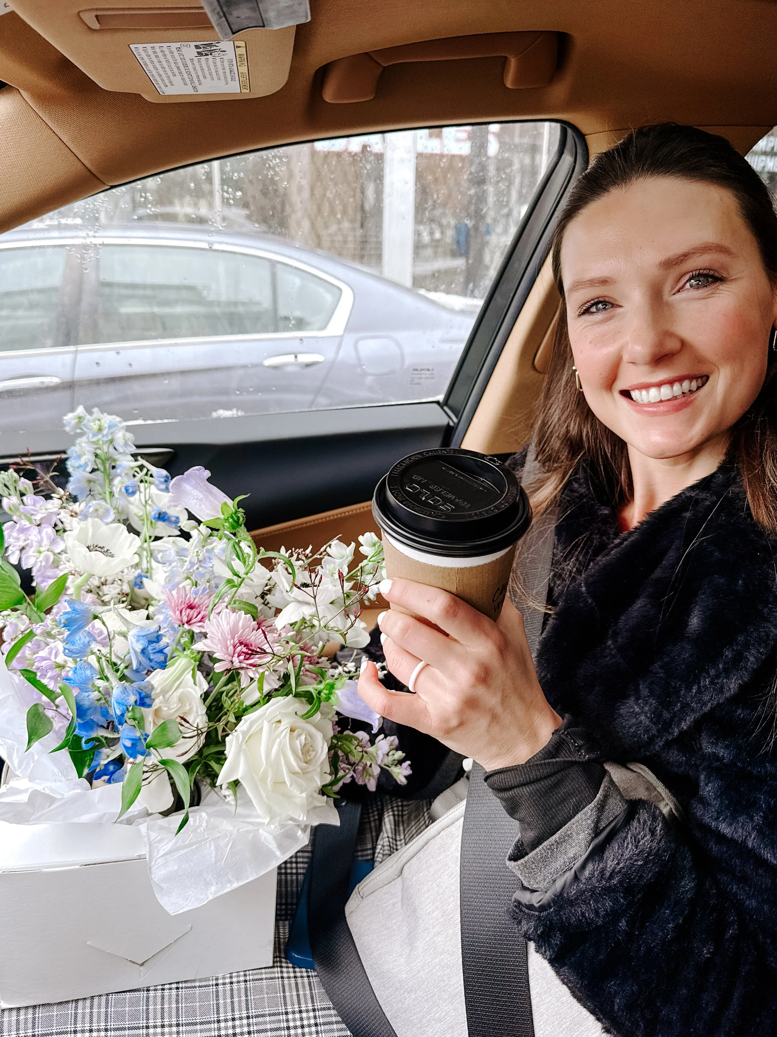 A woman sitting in a car seat, smiling, holding a coffee cup, with a large bouquet of flowers next to her, visible through the car window.