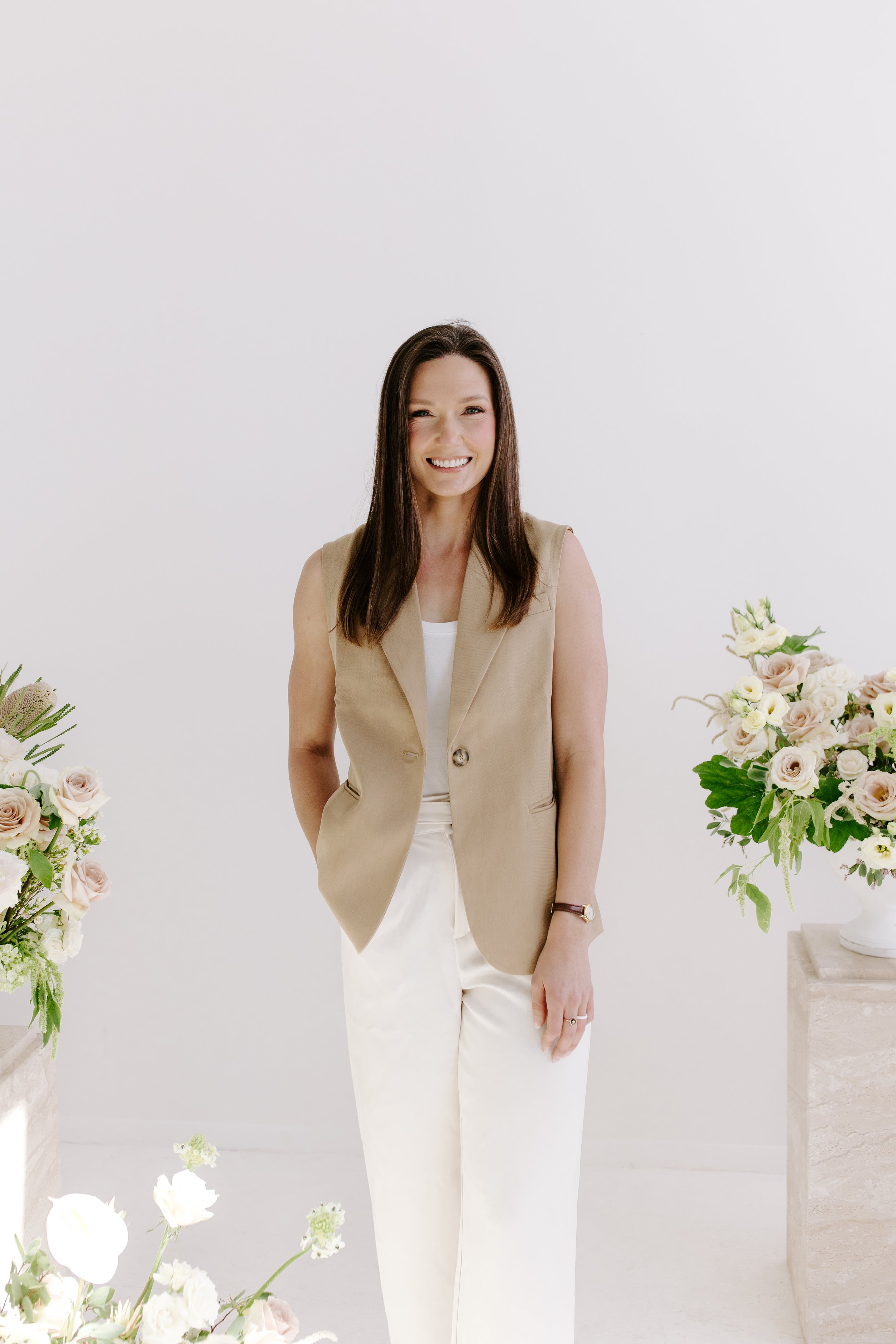 A woman smiling, standing in a white room with floral arrangements on either side.