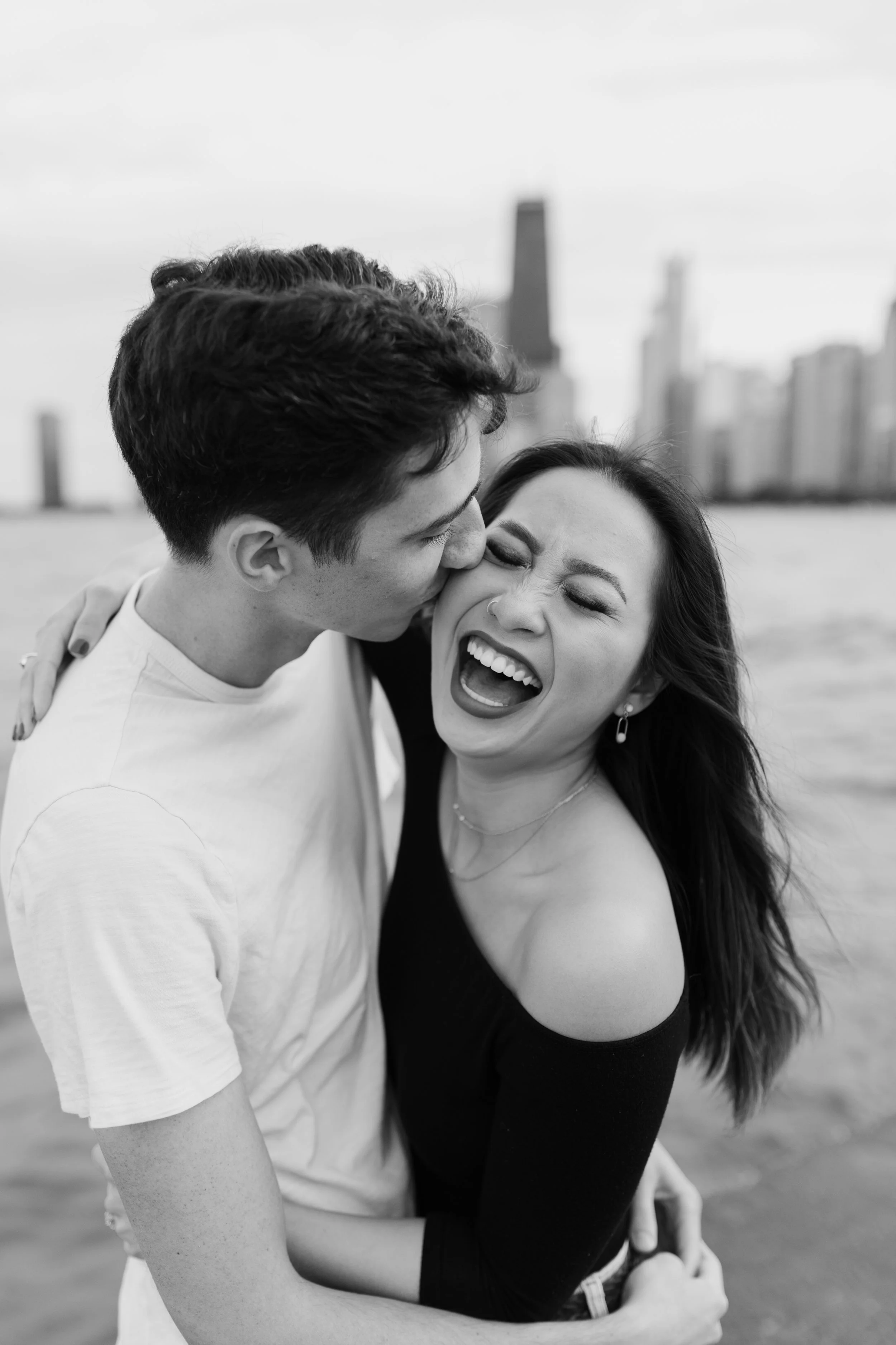 A young man and woman are standing close together near water, with the city skyline in the background. The man is kissing the woman on her cheek while she is laughing with her eyes closed, wearing a black off-shoulder top.