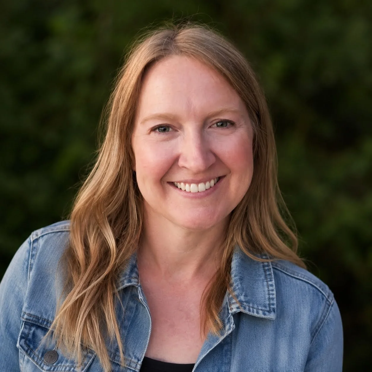 A woman with long, wavy, light brown hair smiling outdoors, wearing a denim jacket and a black top against a blurred green background.
