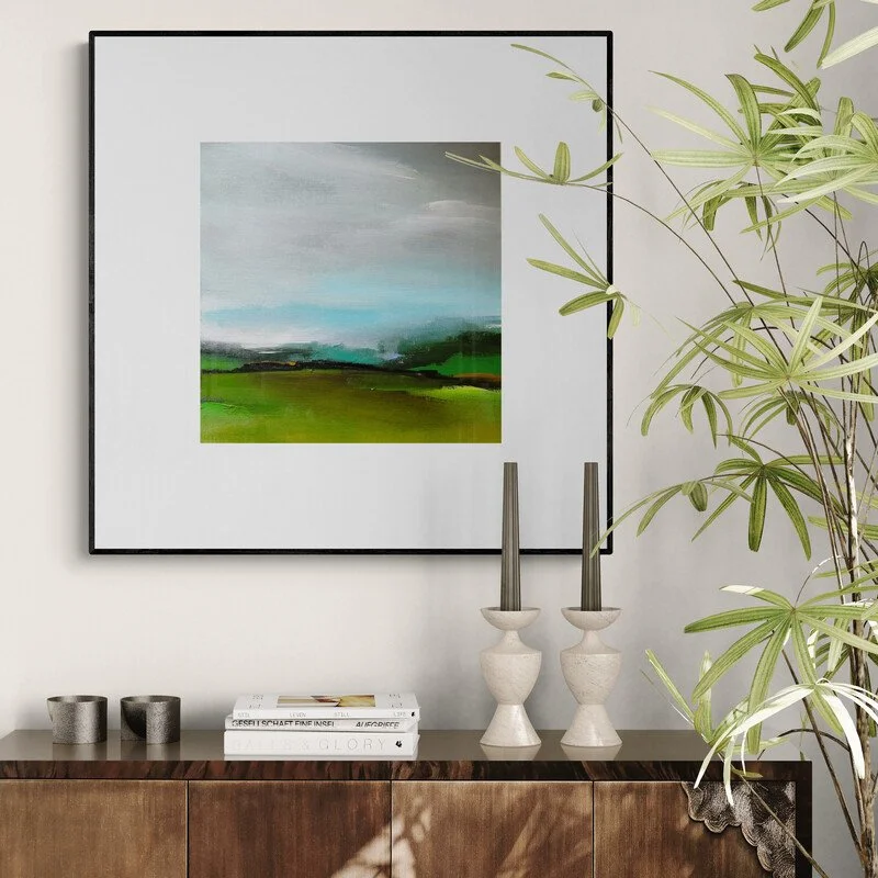 Small landscape painting on canvas framed hanging above plant and candle sticks
