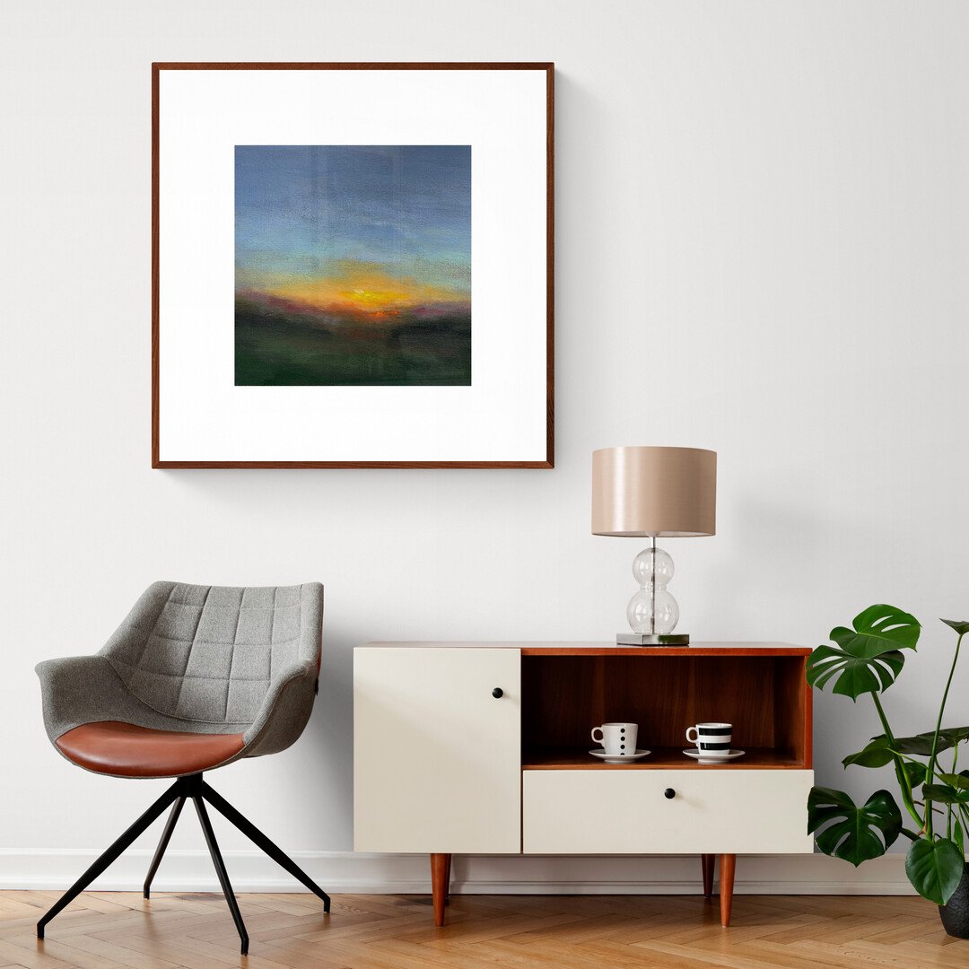 Abstract sunset painting framed and hanging in modern living room above chair, small cabinet and lamp.