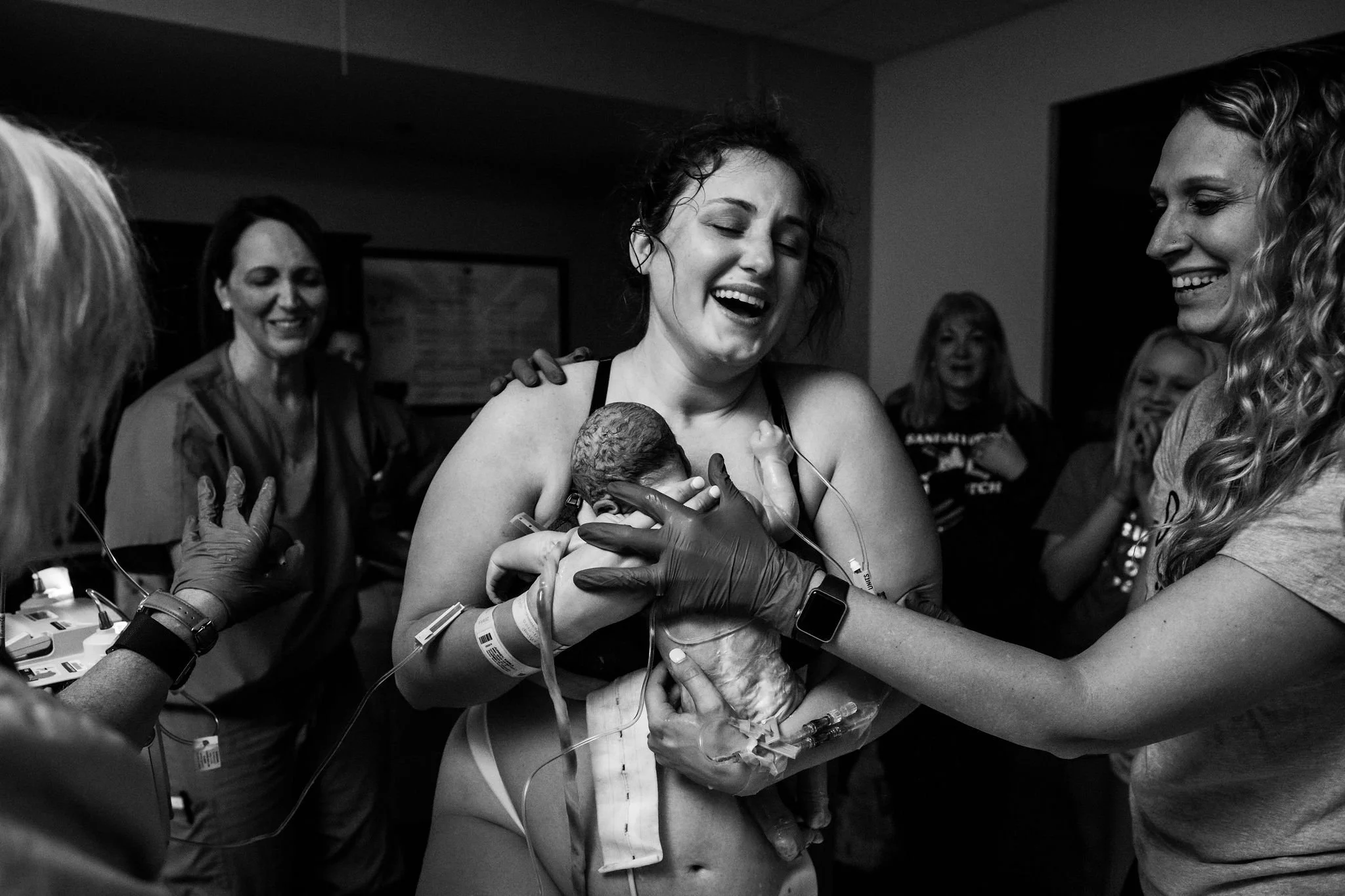 Mother holding newborn baby in hospital surrounded by medical staff and family, expressing joy and emotion.